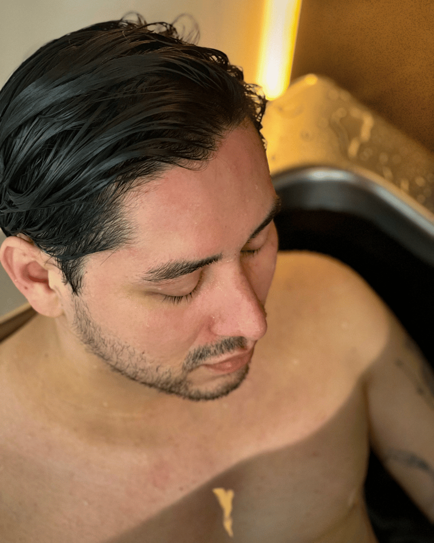 Man with dark hair and a short beard relaxing in a bathtub, eyes closed and water droplets on his shoulder.