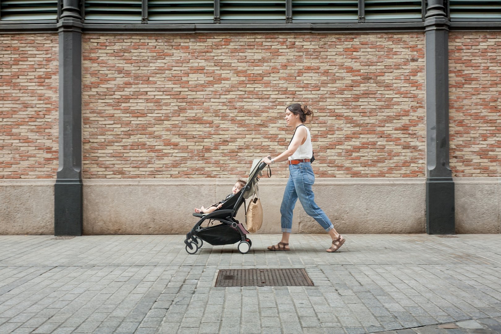 A mother pushes a stroller along a city sidewalk, walking past a patterned brick wall. The side view captures urban texture and everyday parenting in motion.