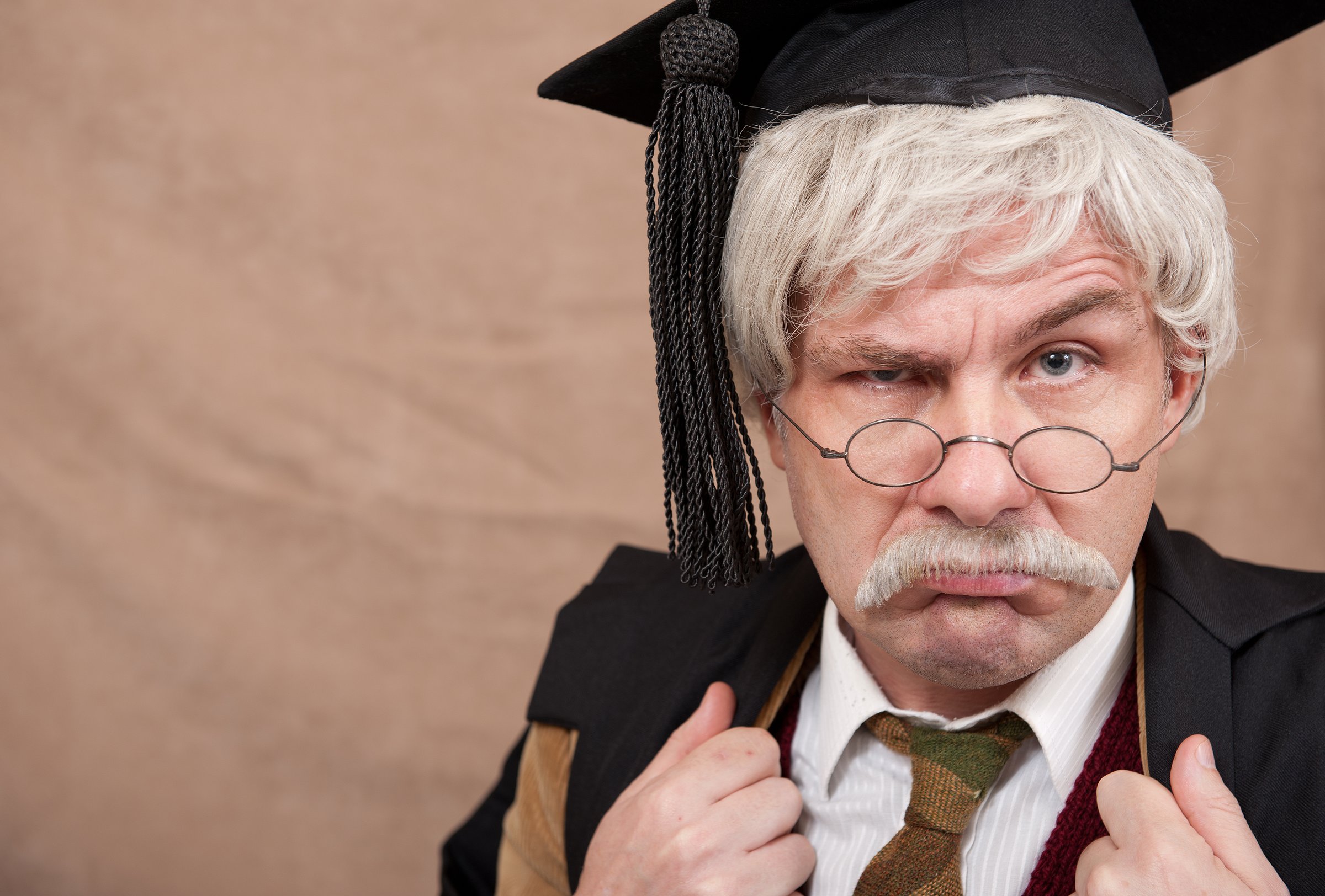 Portrait of an old style English school headmaster with a stern expression, against a light brown background.