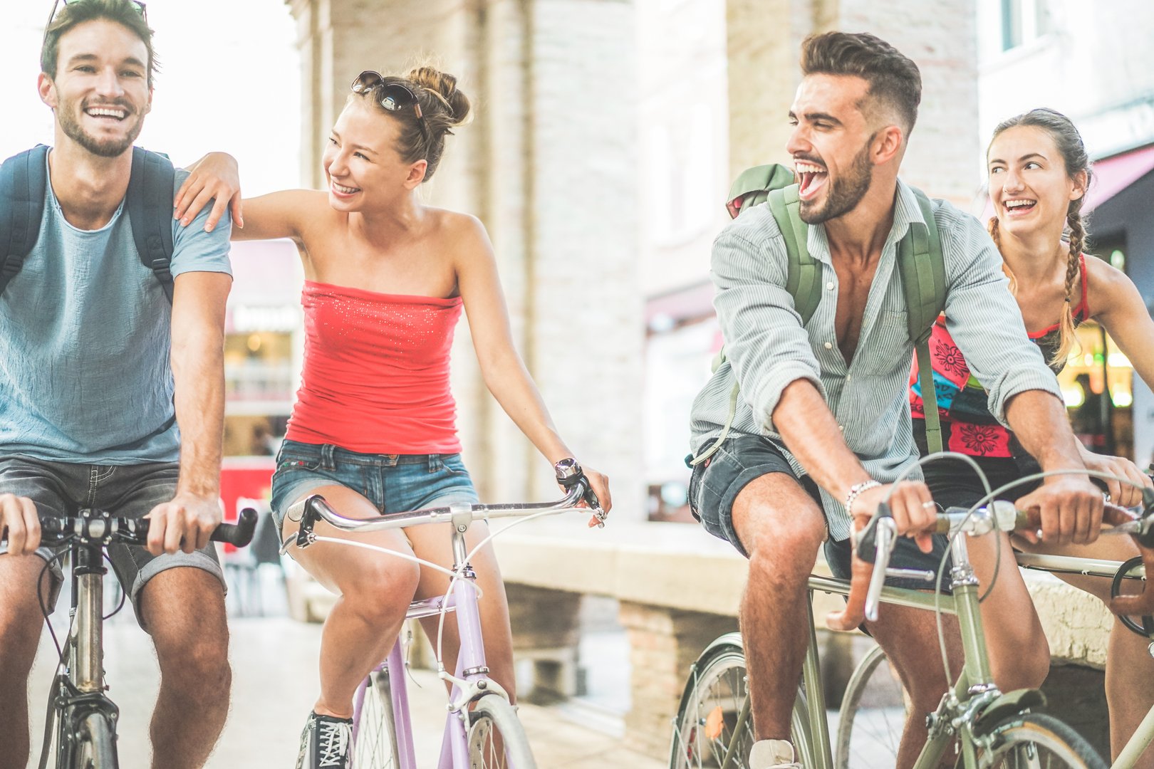 Happy best friends riding bicycles in city center - Young students having fun together around the town - Youth and friendship concept - Soft focus on left woman face - Warm filter