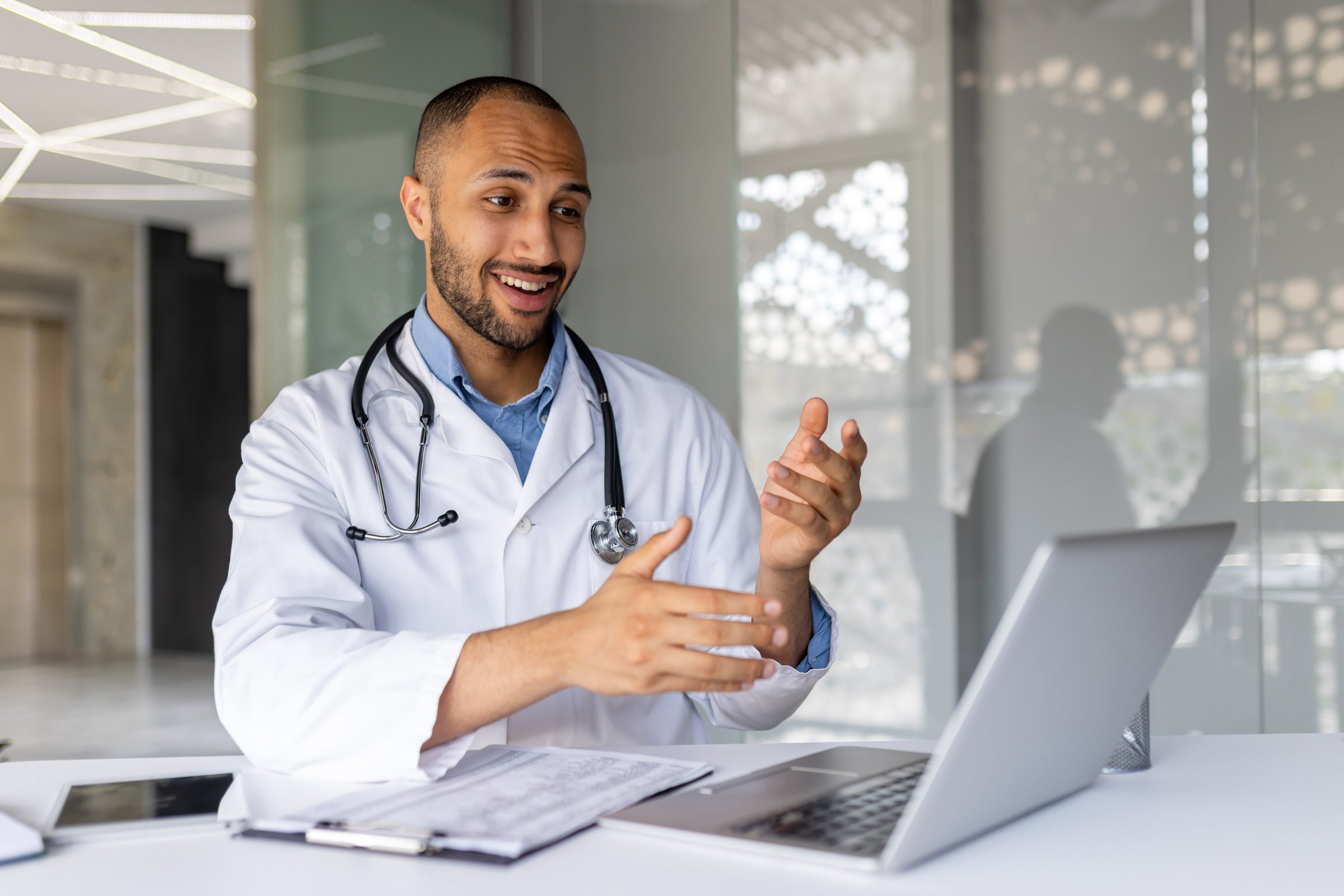 Smiling doctor with stethoscope uses laptop for virtual consultation in modern office. Engages in telemedicine call, representing efficient healthcare technology and professional communication.