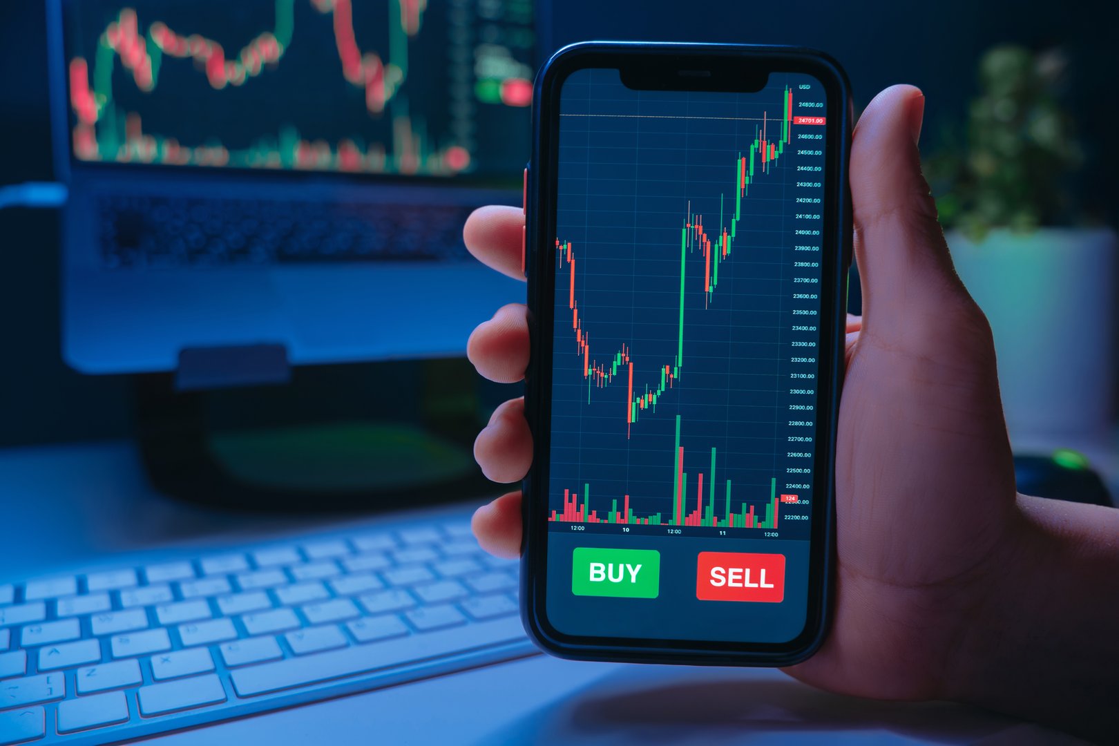 Trader hand holding smartphone with stock market or crypto trading app on screen with buy and sell buttons, against desk with laptop. Online cryptocurrency trading and investment. Space for text