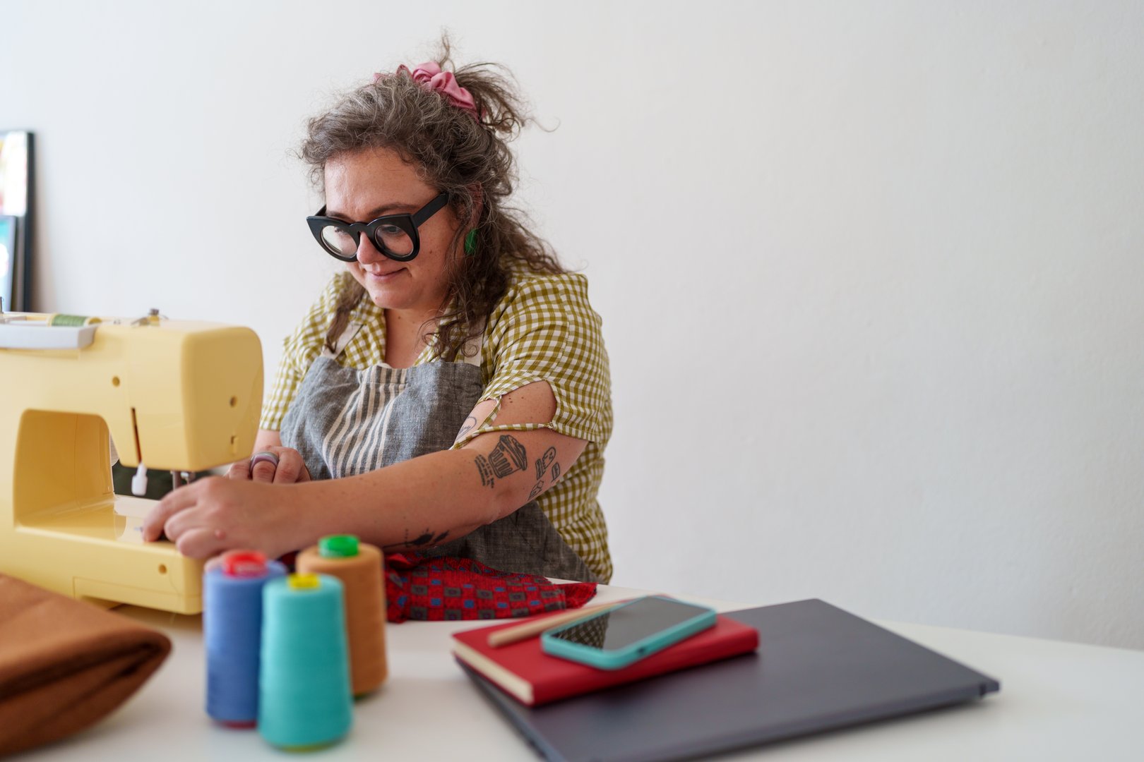 Tailor working with a sewing machine, colorful thread spools and fabrics on the foreground, notebook and smartphone on a white table