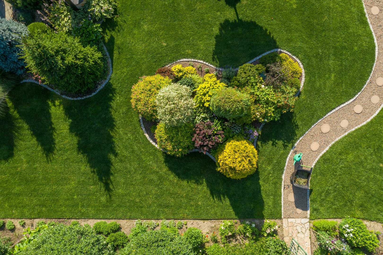 Garden Maintenance and Care Work in Process. Wheelbarrow with Grass Cuttings and a Plants Watering Can Stand on a Twisting Garden Path Next to a Large Landscape Island. Aerial View.