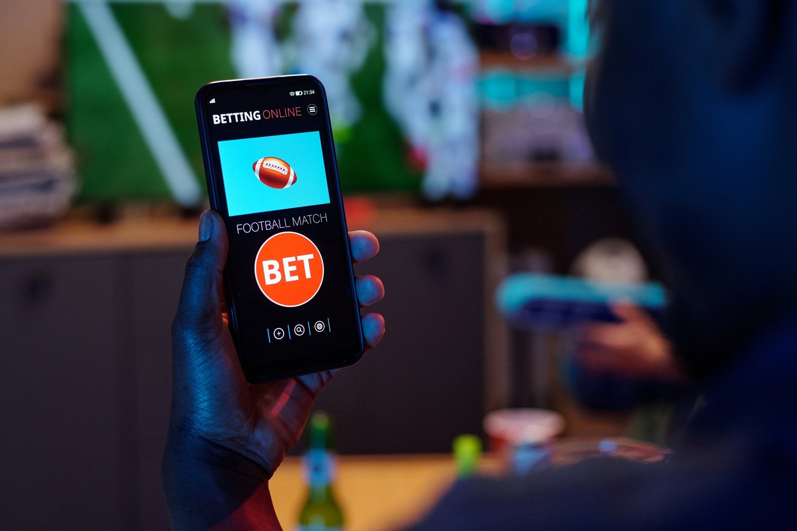 Black man holding smartphone displaying online sports betting app with football match bet option visible on screen, sitting indoors with blurred background and beverage bottle