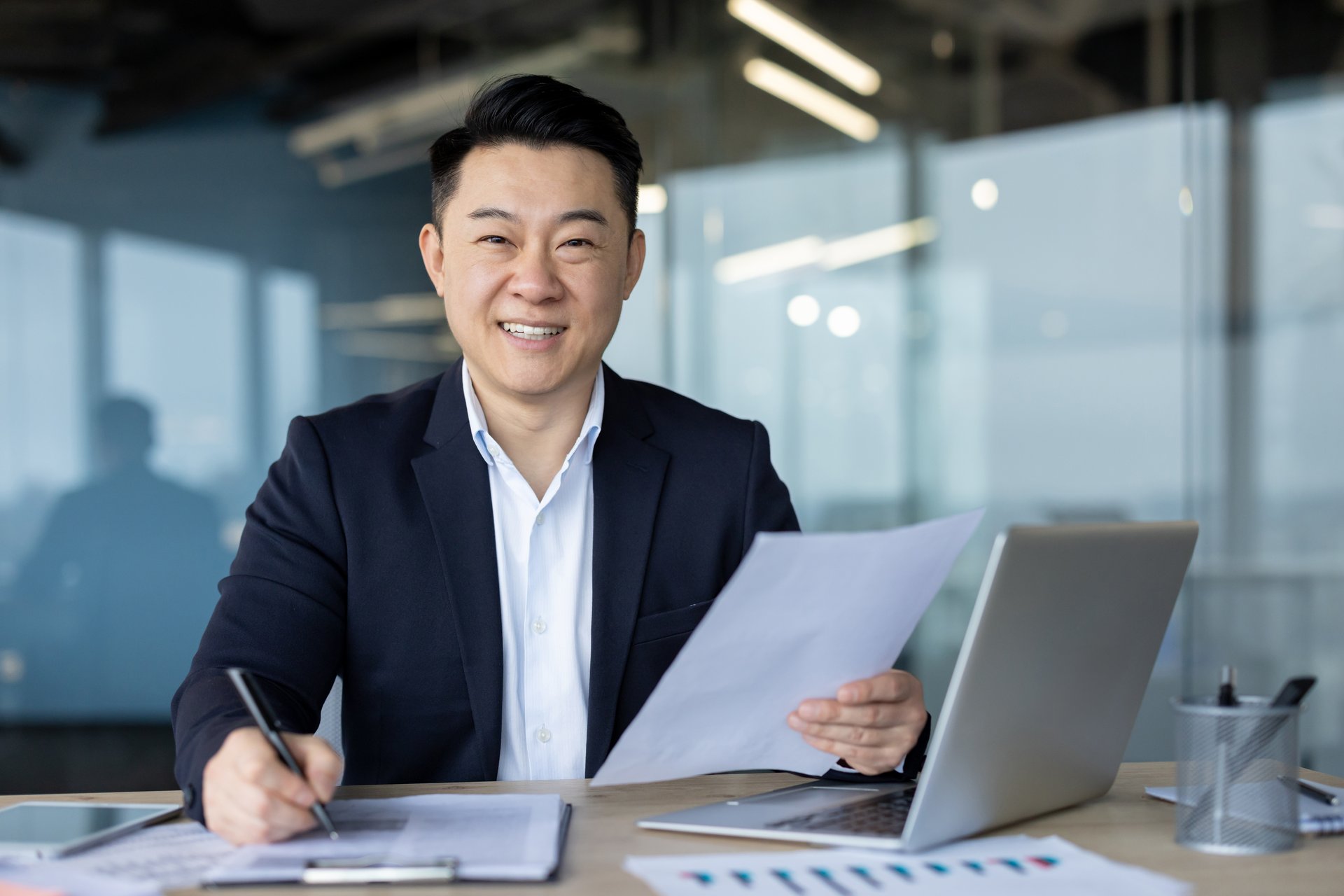 Portrait of a successful young Asian businessman working in the office with a laptop and documents, smiling and looking at the camera.