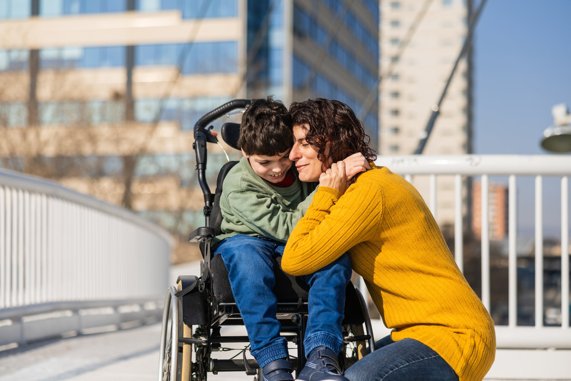 Mother lovingly embraces her son with disability  who is sitting in a wheelchair, showing affection and support on a bridge in the city