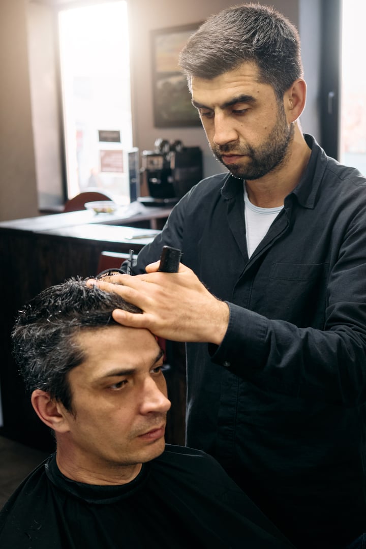 A hairdresser expertly cuts a customer's hair in a contemporary barbershop. Natural light fills the space, enhancing the professional atmosphere of the haircutting session.
