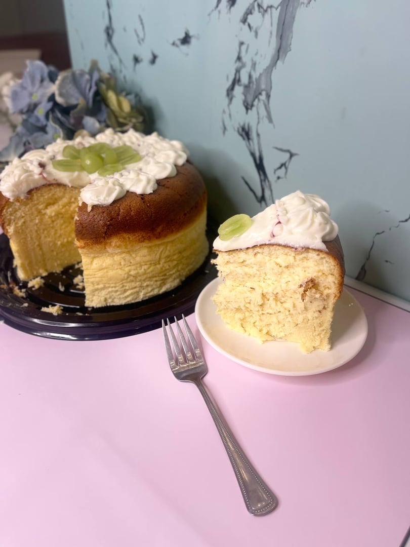 A slice of fluffy cake with cream and grapes on top, placed on a plate next to the rest of the cake and a fork.