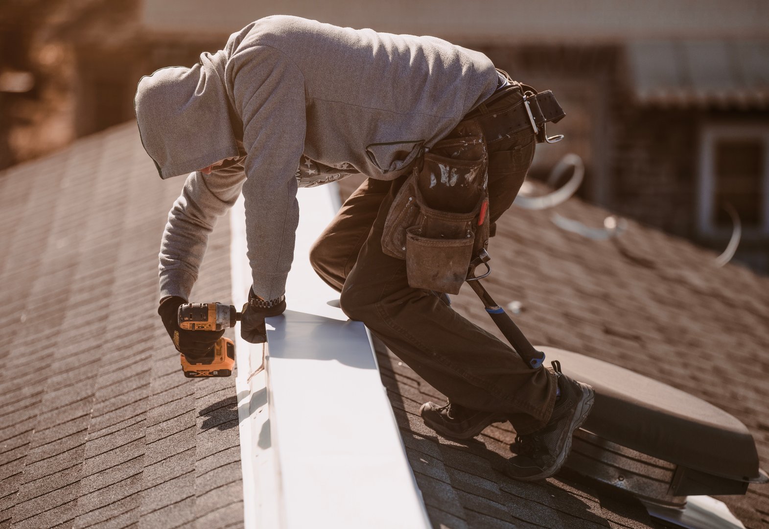 A roof repairman installs a metal ridge cap on top of a roof to seal and protect the roofline during maintenance. He is working with tools and safety gear under clear daylight, ensuring the roof's durability and weather resistance.