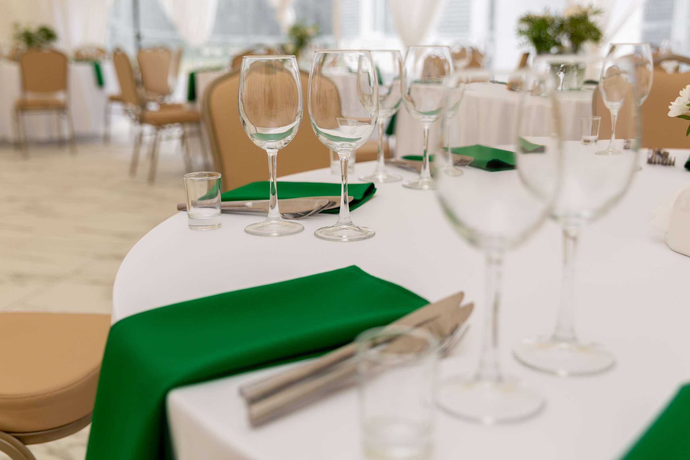 Close up of wine glasses, silver tableware and green napkins on round tables covered with white tablecloth. Catering concept. Interior of tent for wedding dinner, ready for guests.