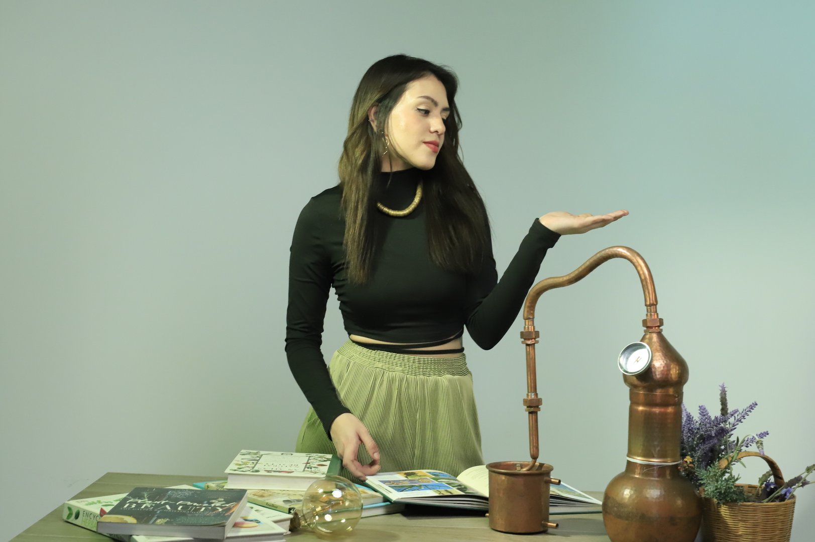 Woman standing by a table with books and a copper distiller, gesturing with one hand. She wears a black top and green skirt.