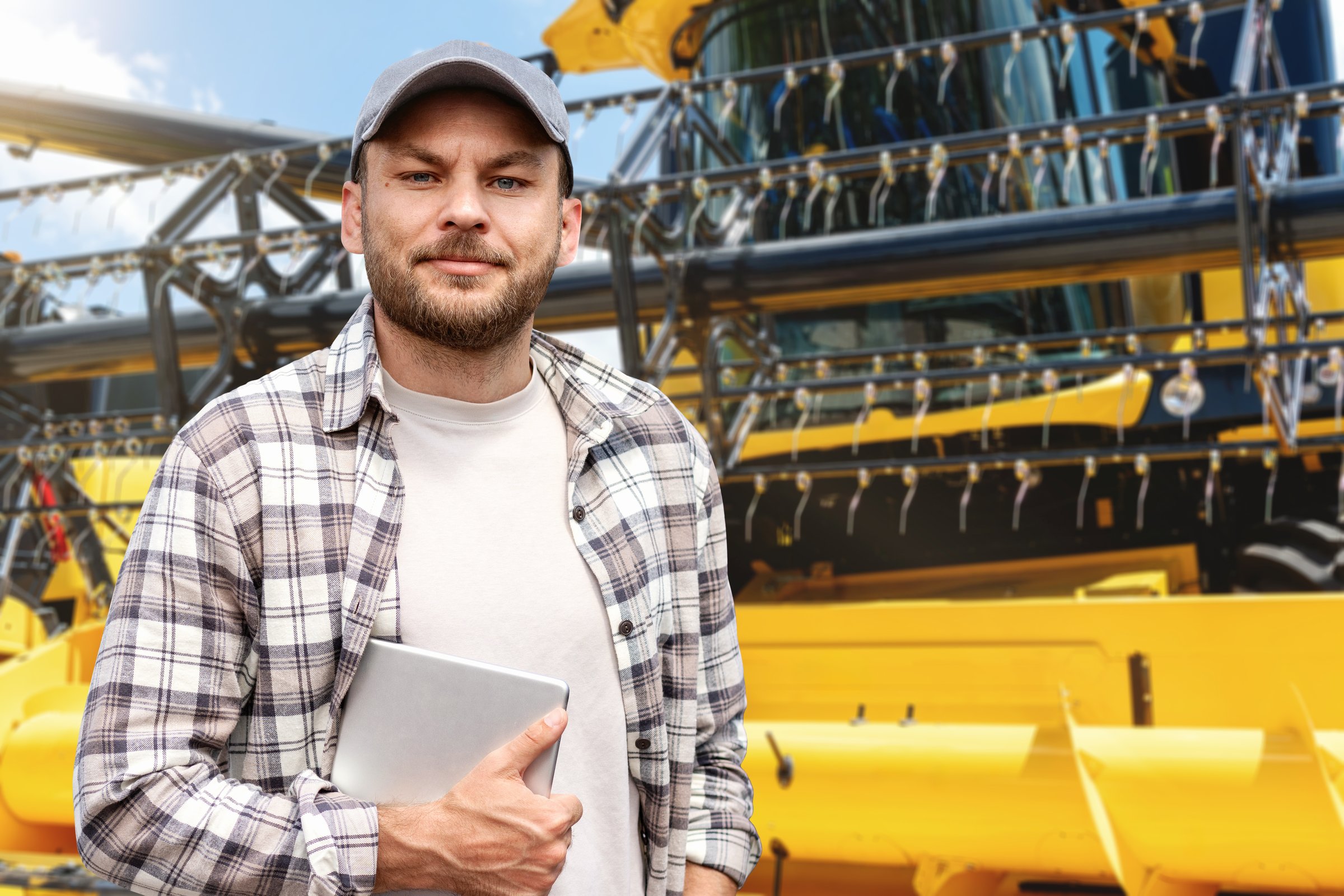 Smiling adult man with digital tablet stands in front of yellow combine harvester outdoors.