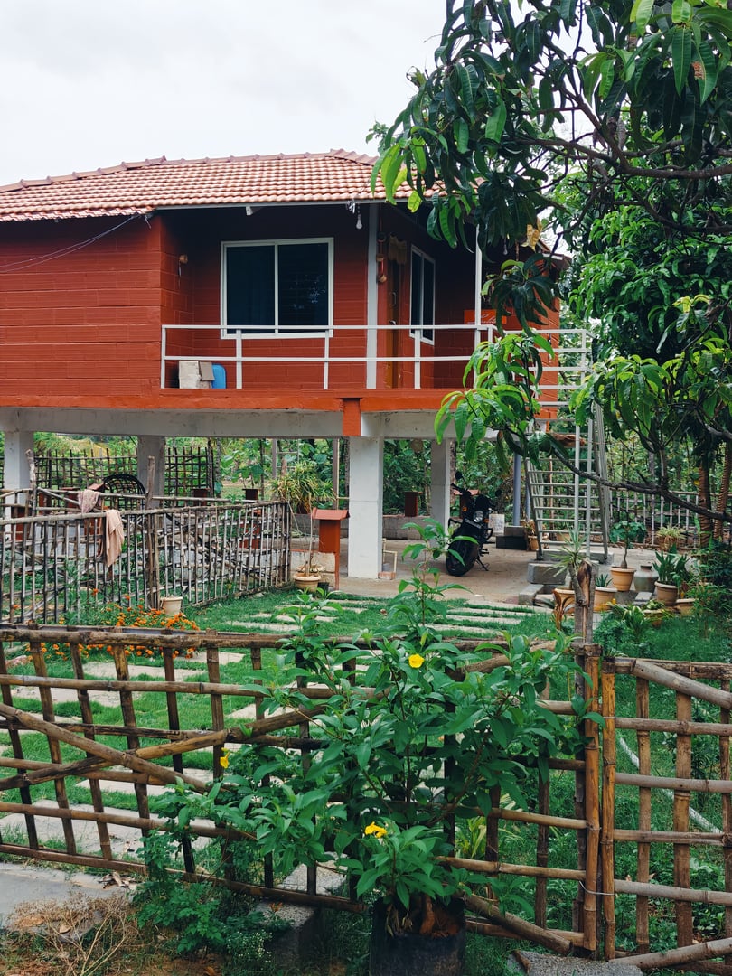 A raised red house with a tiled roof, surrounded by greenery and a wooden fence, with potted plants and a ladder nearby.