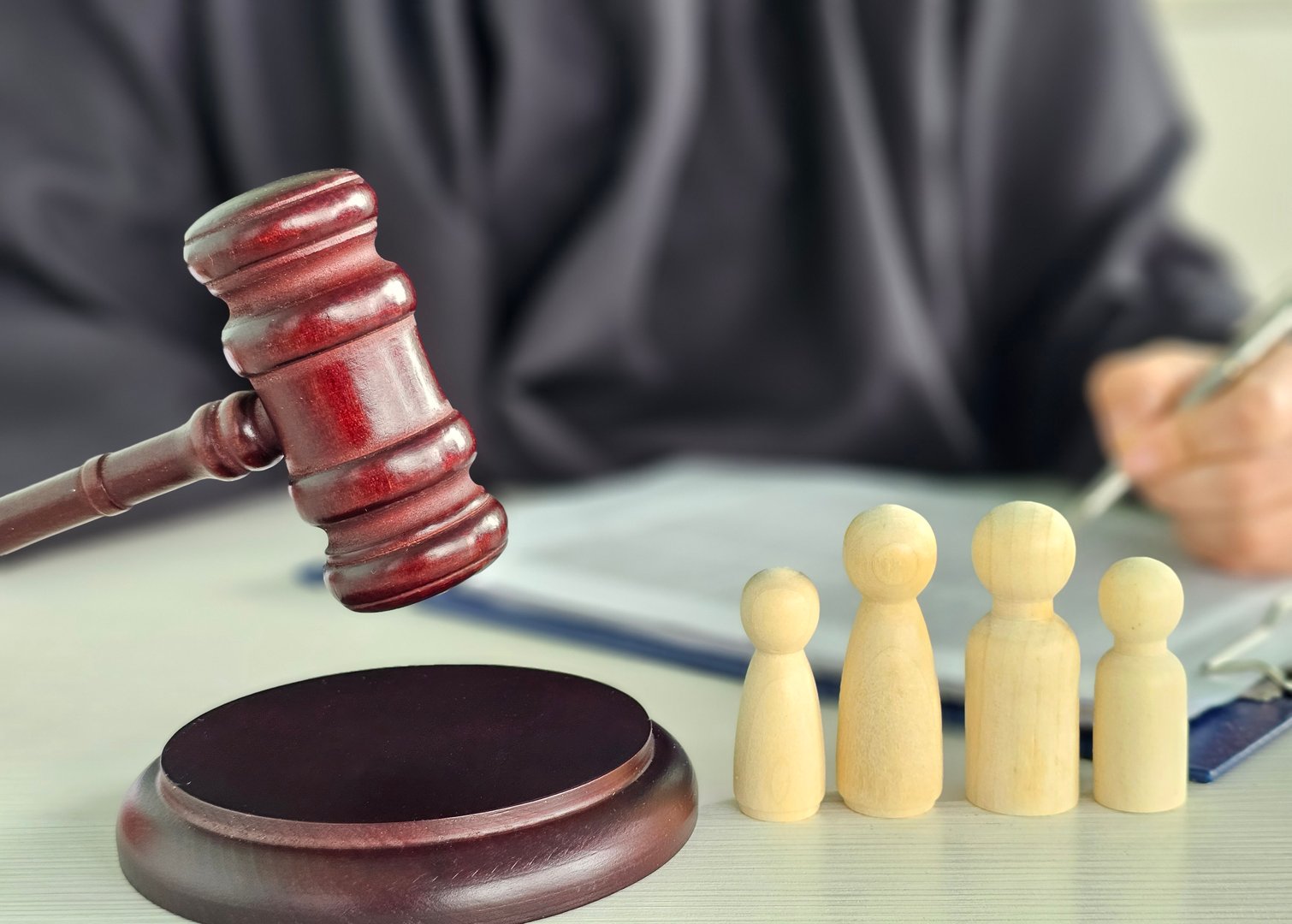 Gavel, sound block and little wooden figures of parents and children placed on desk in courthouse up close, judge and scales of justice in background. Family law, court, divorce, child custody concept