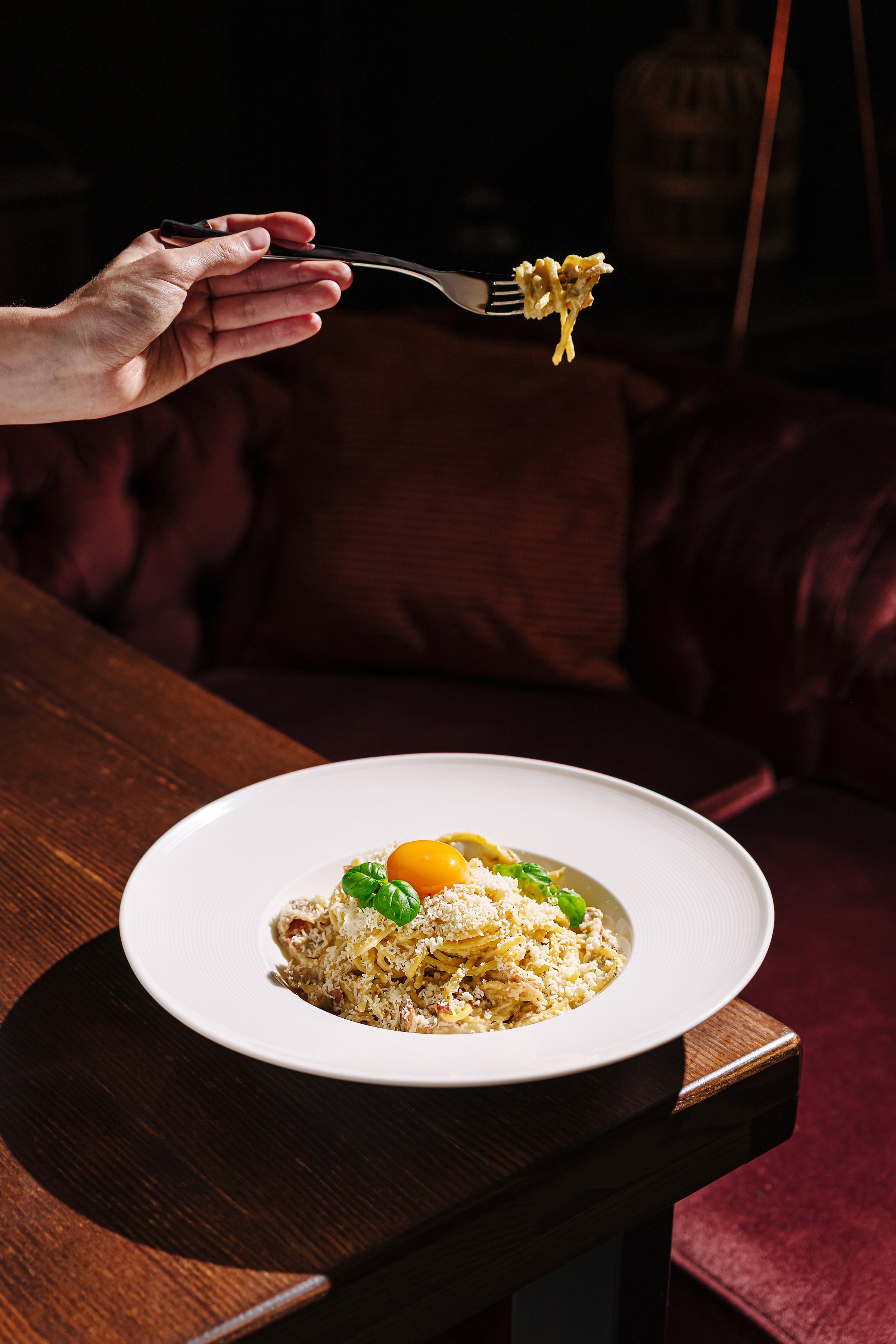 A hand reaches for a forkful of flavorful pasta topped with colorful vegetables and a bright egg yolk on a white plate. The dimly lit restaurant offers a cozy atmosphere.
