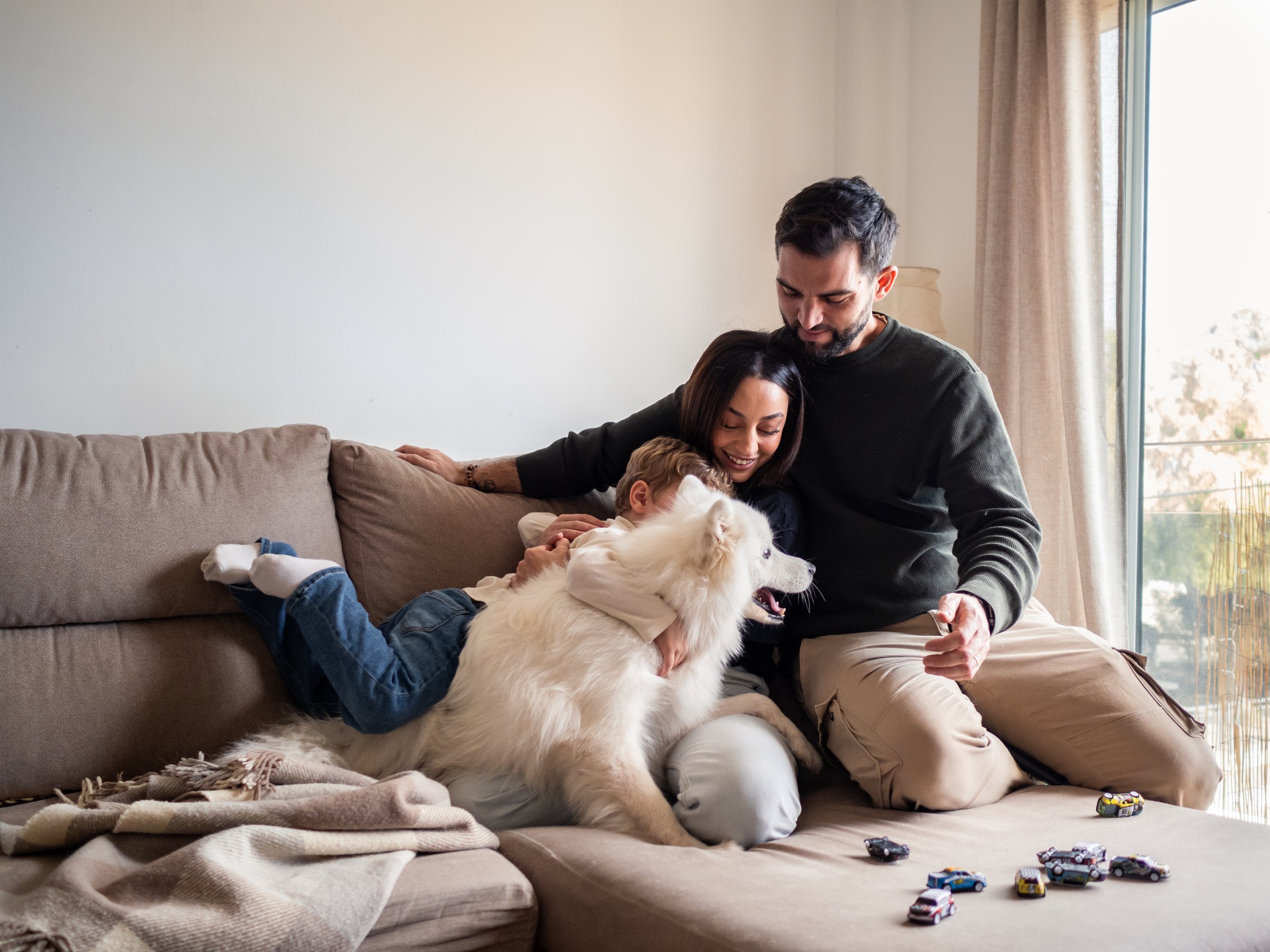 A multiracial young family has a joyful time together on a sofa. The child cuddles with their dog while the parents share smiles, creating a warm atmosphere in their modern living room.