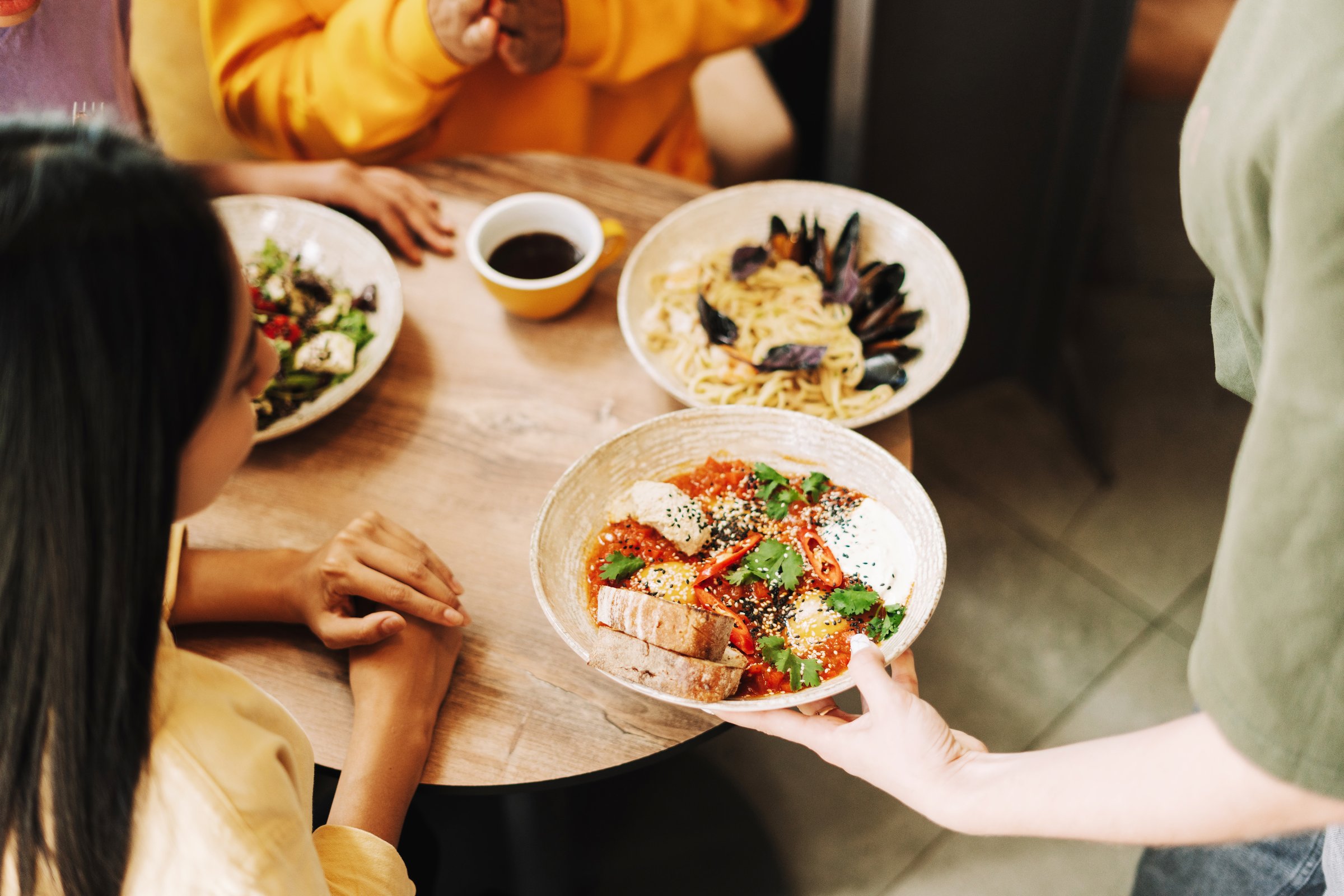 Waitress is holding a plate of dish, serving it to a customer in a restaurant. There are other plates on the table with different dishes