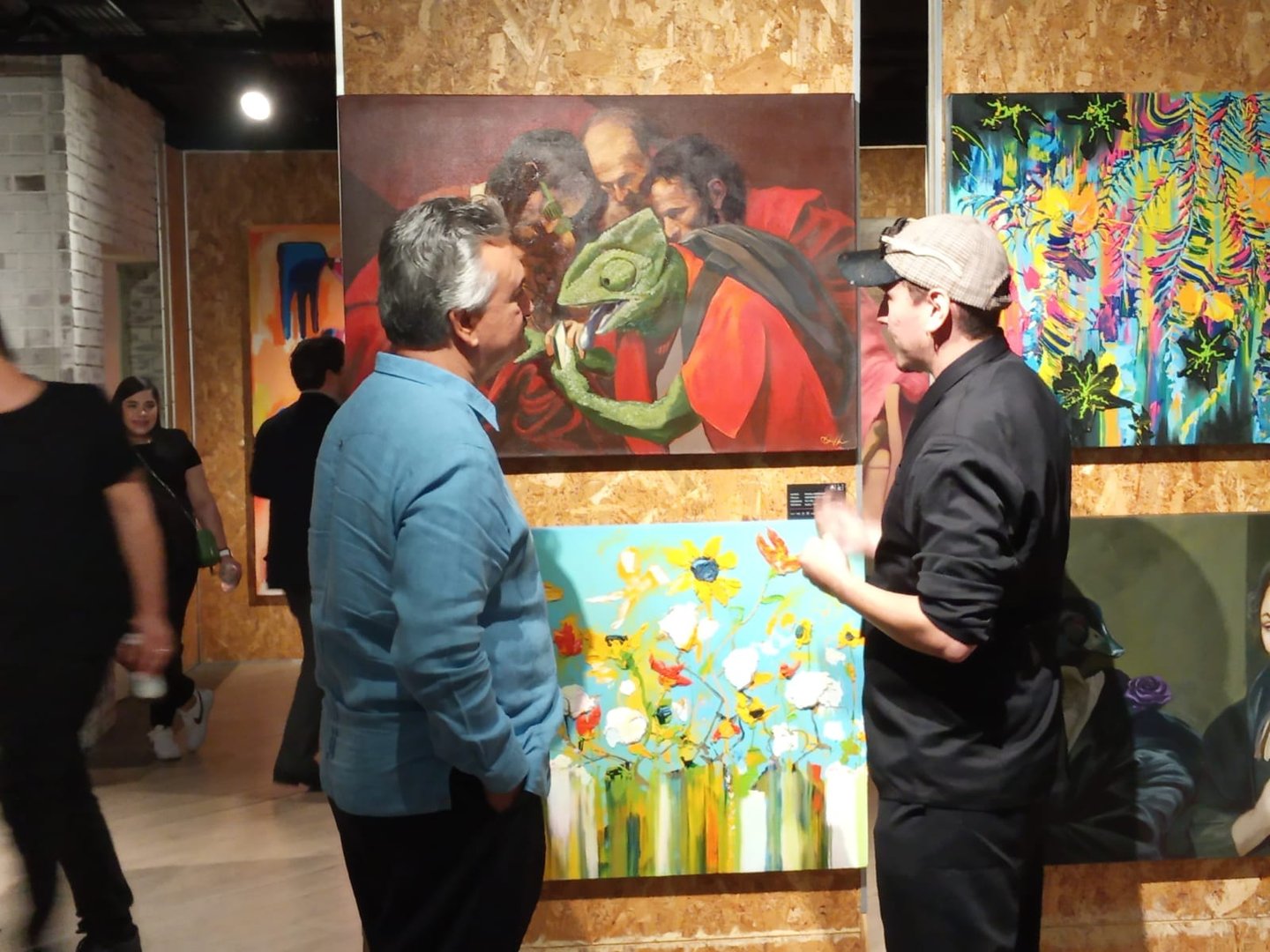 Two men talking in an art gallery, with colorful paintings on the walls, including a lizard-themed artwork.