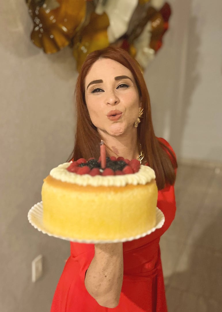 Woman in a red dress holding a cake with fruit toppings and a lit candle, posing in a celebratory manner indoors.