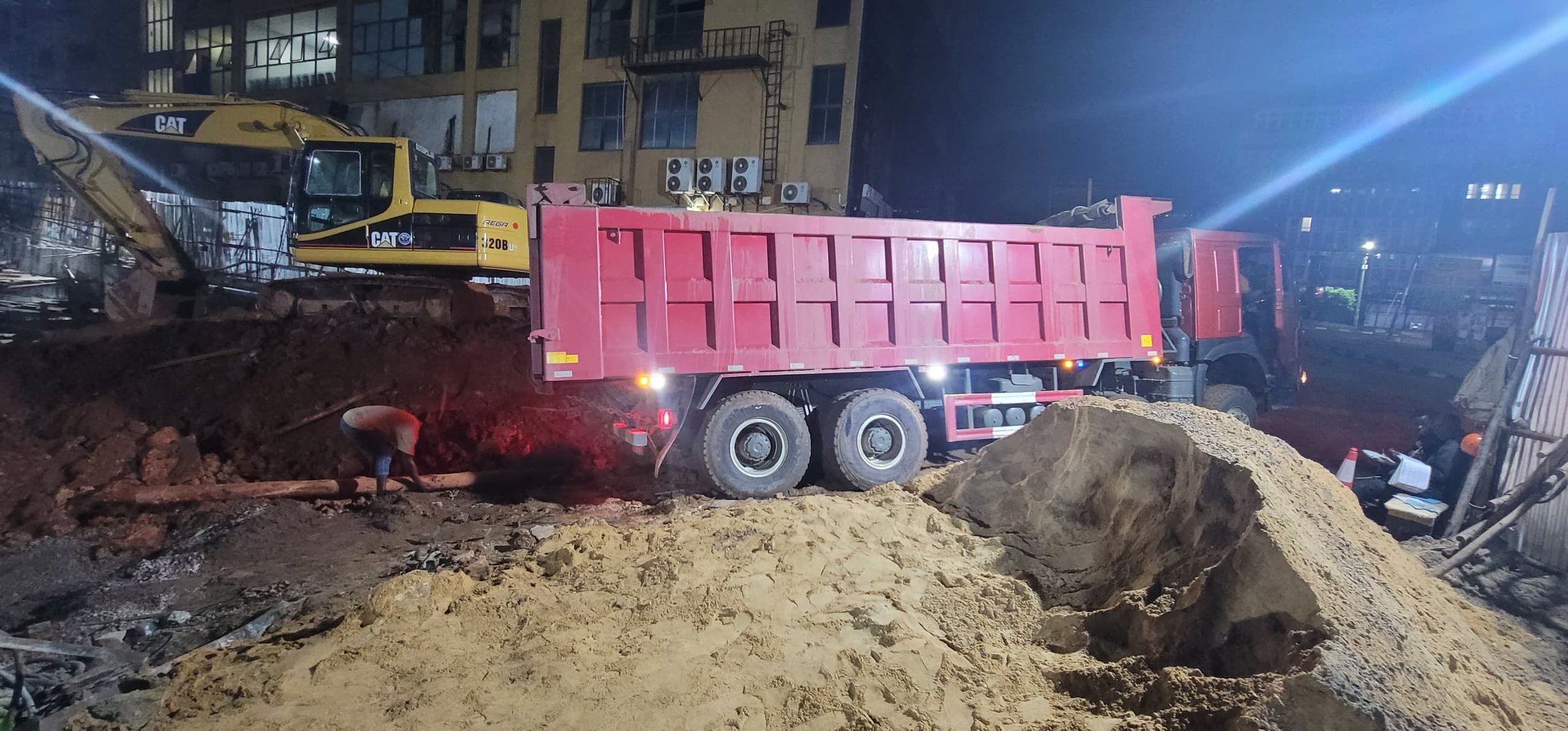 Nighttime construction site with a red dump truck parked on sandy terrain, surrounded by heavy machinery and workers.