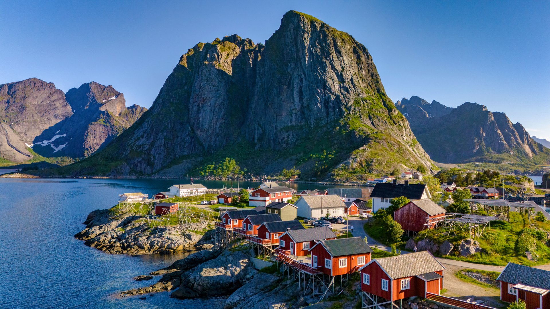 A picturesque view of a small village in Norway, featuring red cabins nestled among rugged mountains and a calm blue fjord. Hamnoy Lofoten Norway in summer