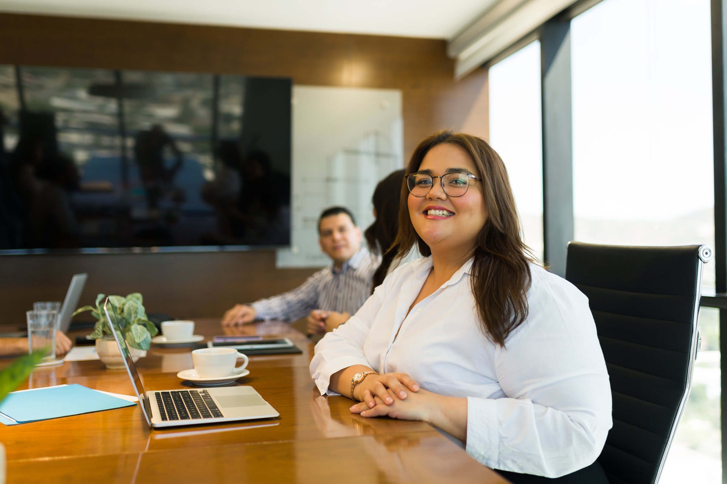 Beautiful smiling plus size businesswoman sitting at meeting room table looking at camera with colleagues discussing in background