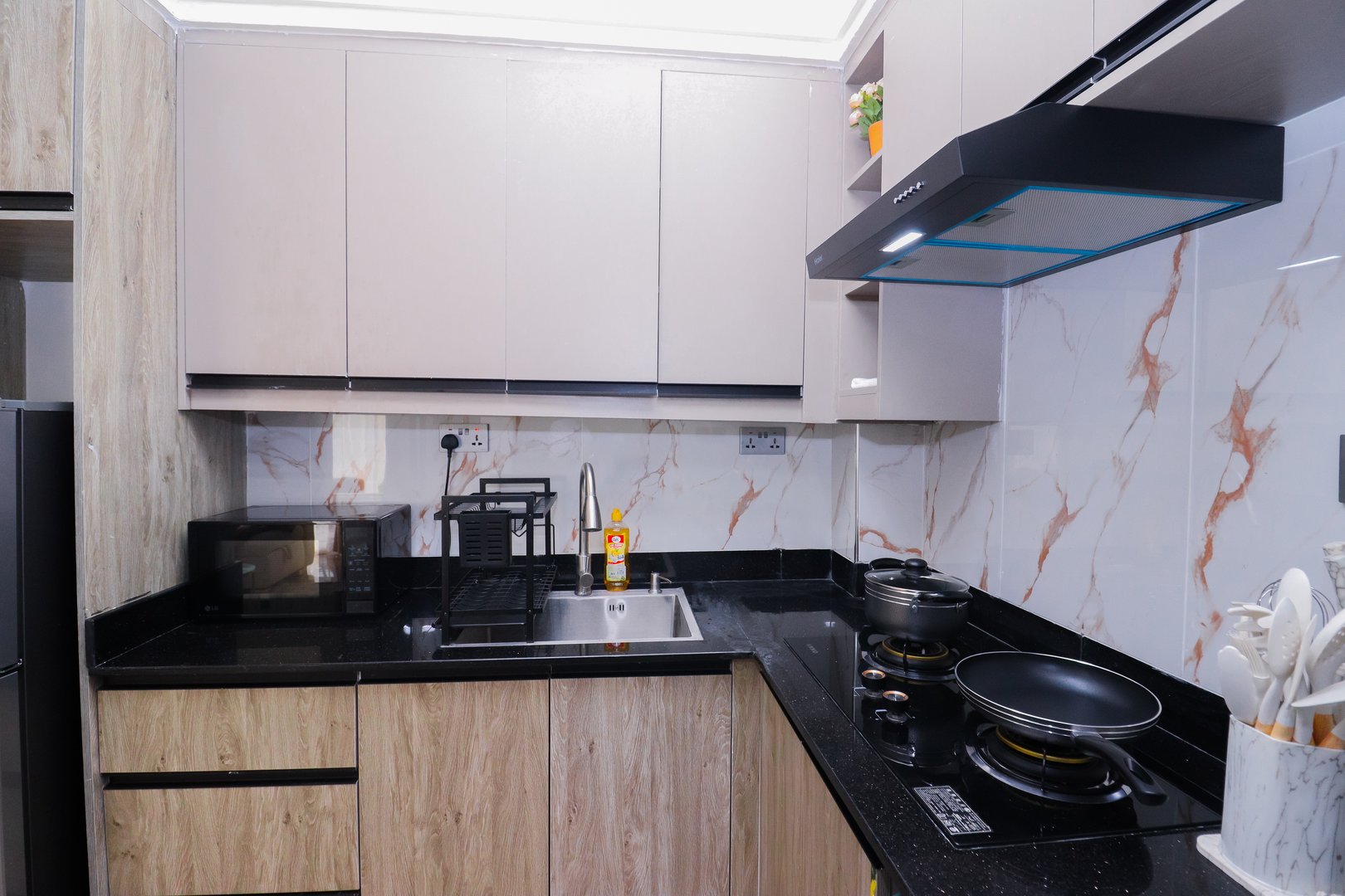 Modern kitchen with black countertops, stainless steel sink, microwave, stove with pans, and wood-tone cabinets.