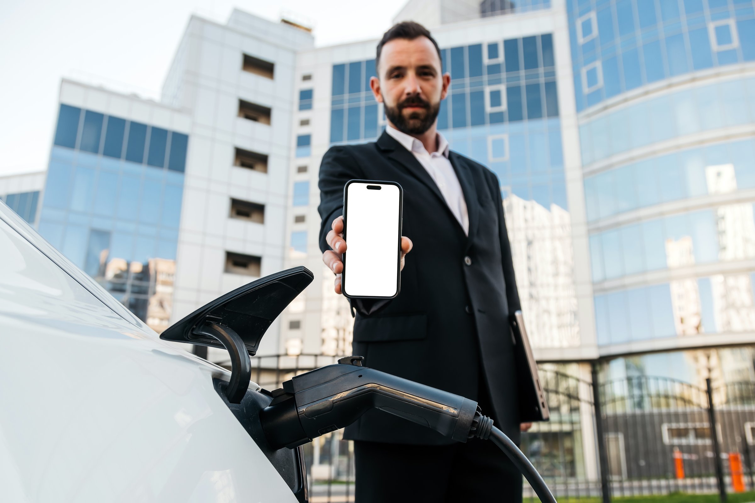 Business man in classic suit near charging station charging his electric car and holding a phone with white screen. Layout for your advertising.
