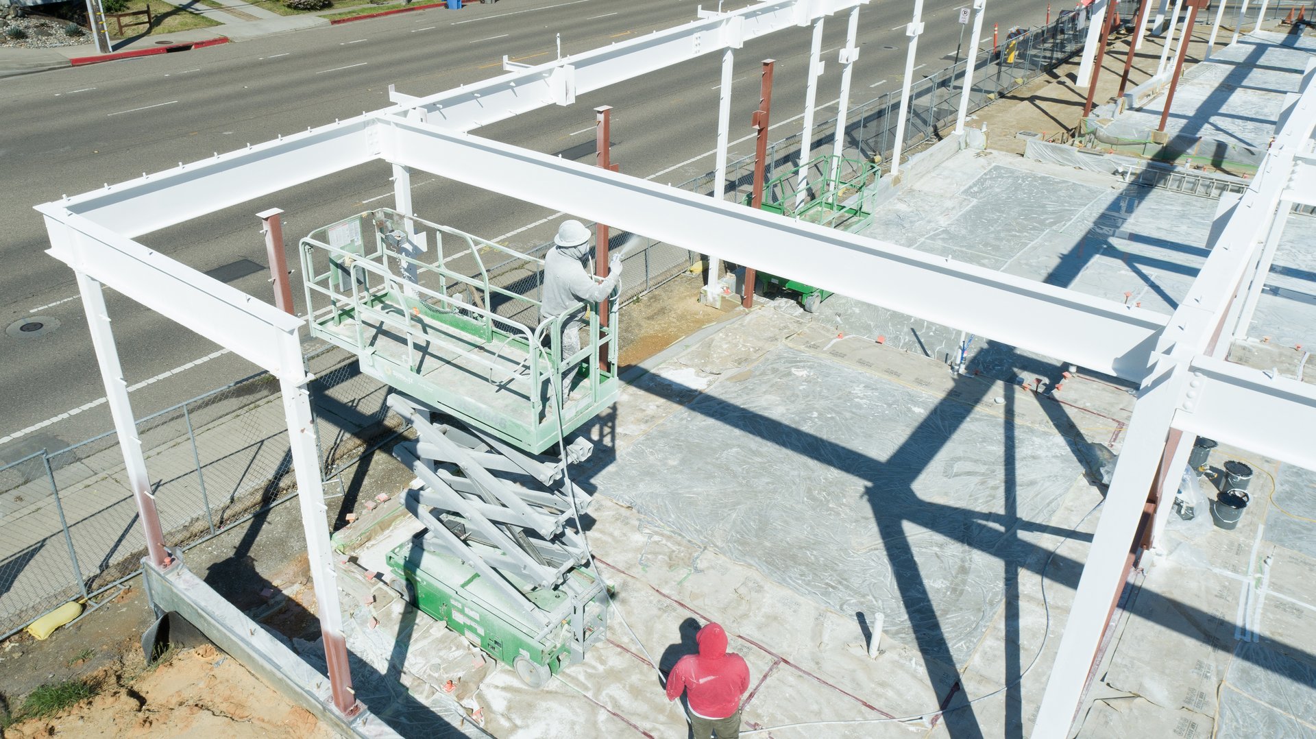 Building construction site with steel beam construction. Man with construction hat and coveralls is standing high up on scissor lift spray painting the beams with white paint. Man in red hoodie stands below observing. Paved road is in the background.