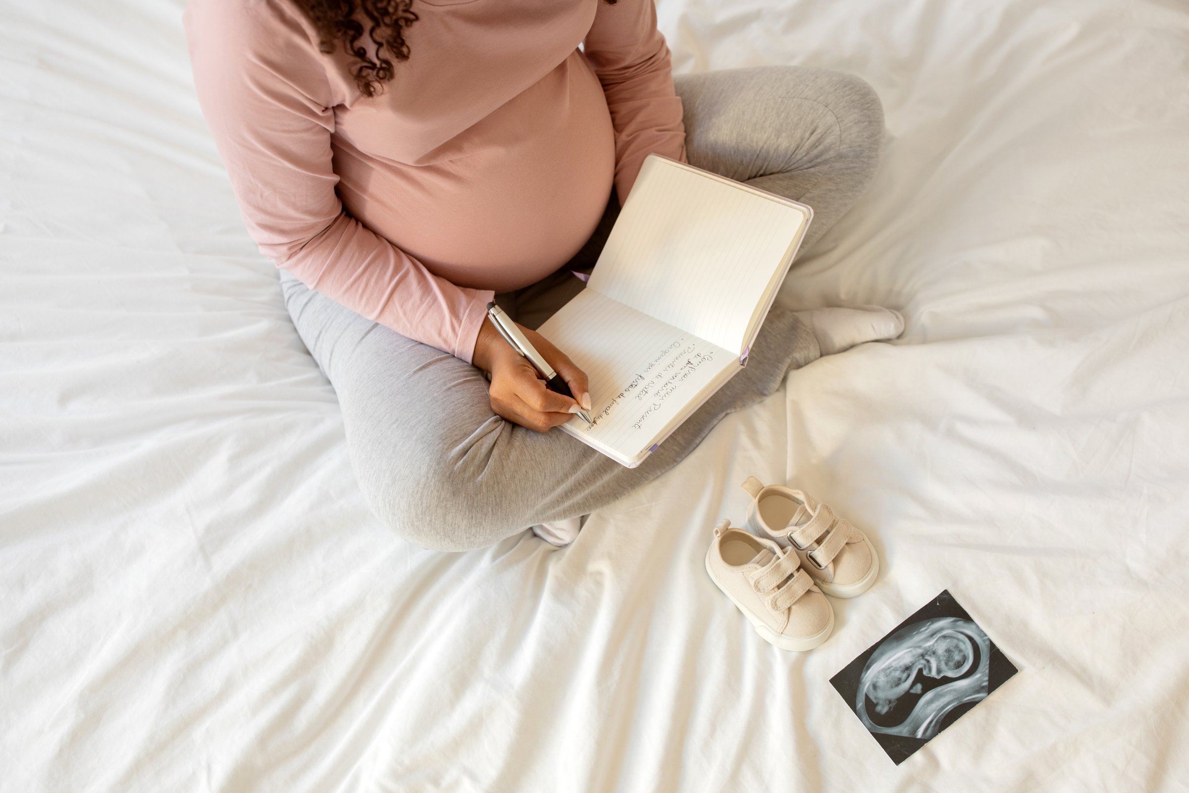 Pregnant woman writing down thoughts in her notebook at home, sitting on bed surrounded by tiny shoes and sonogram picture, expectant mother creating memories during her pregnancy or making checklist