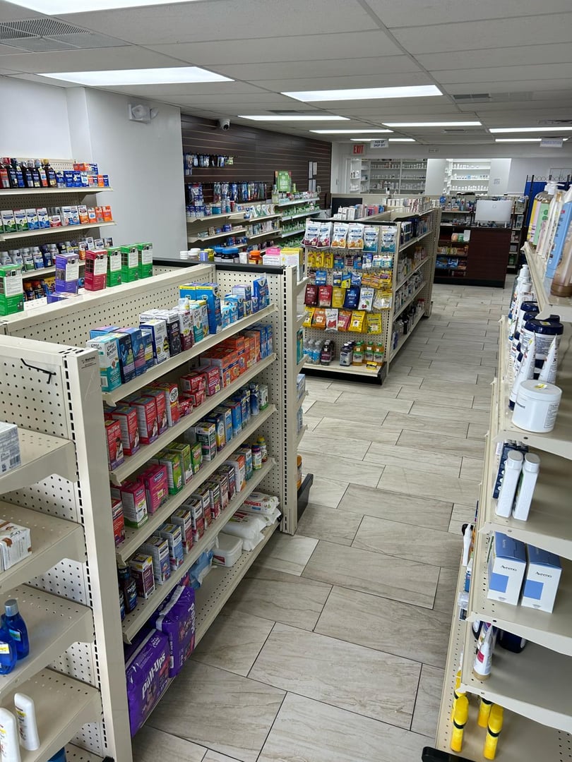 Interior of a pharmacy with shelves stocked with various medications and products, light wood flooring, and bright lighting.
