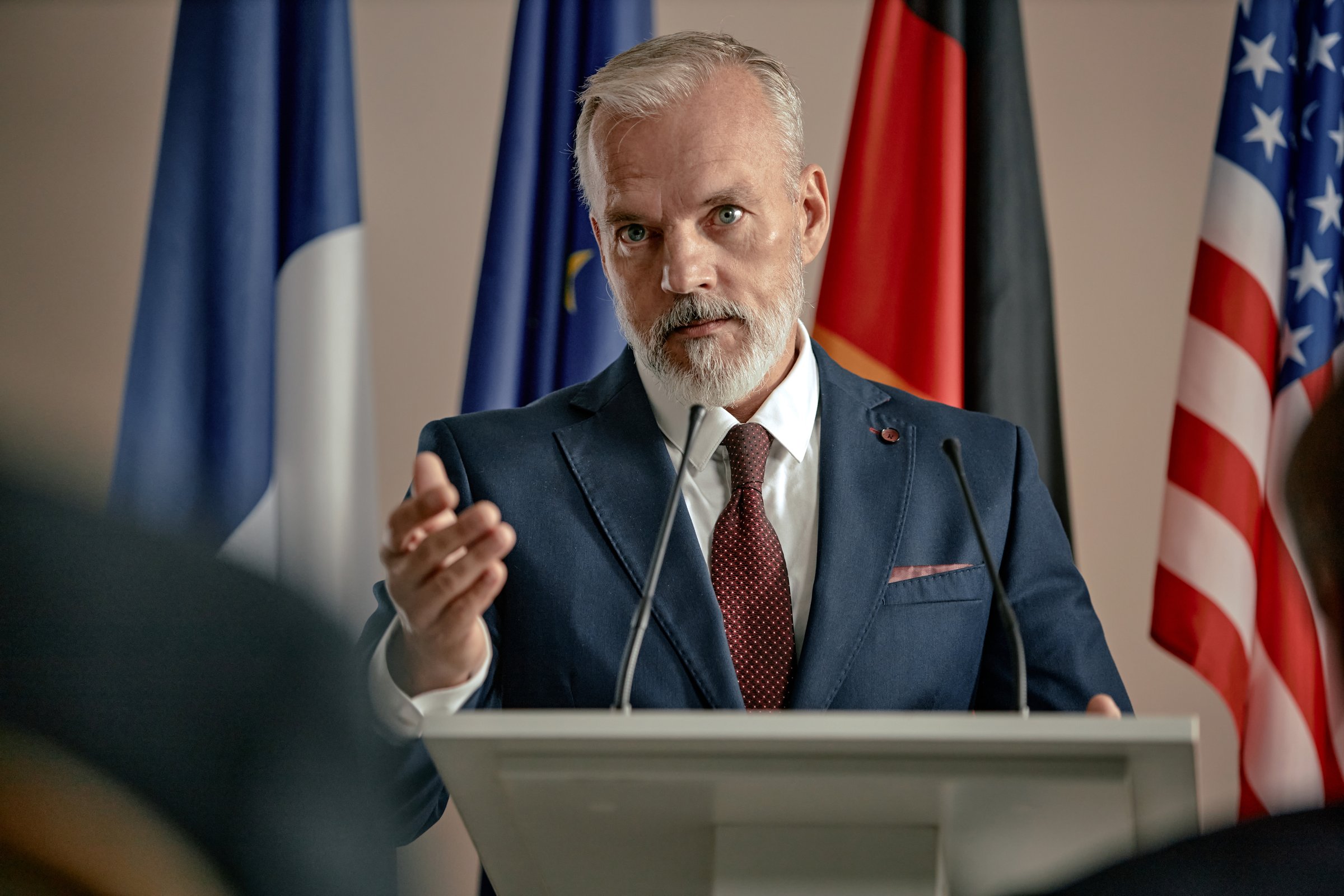 Portrait of senior Caucasian man speaking at podium gesturing with hand during press conference with international flags in background, maintaining serious facial expression