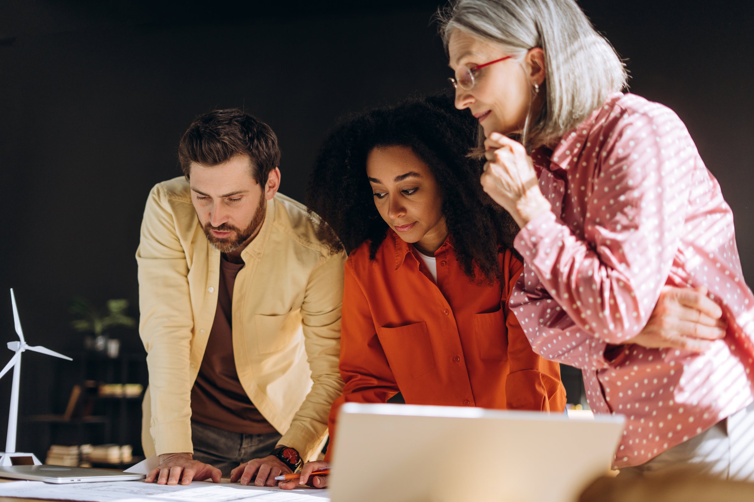 Group of multinational engineers working together on blueprints for sustainable project, using laptop and with wind turbine model on desk, showcasing to renewable energy. Teamwork concept