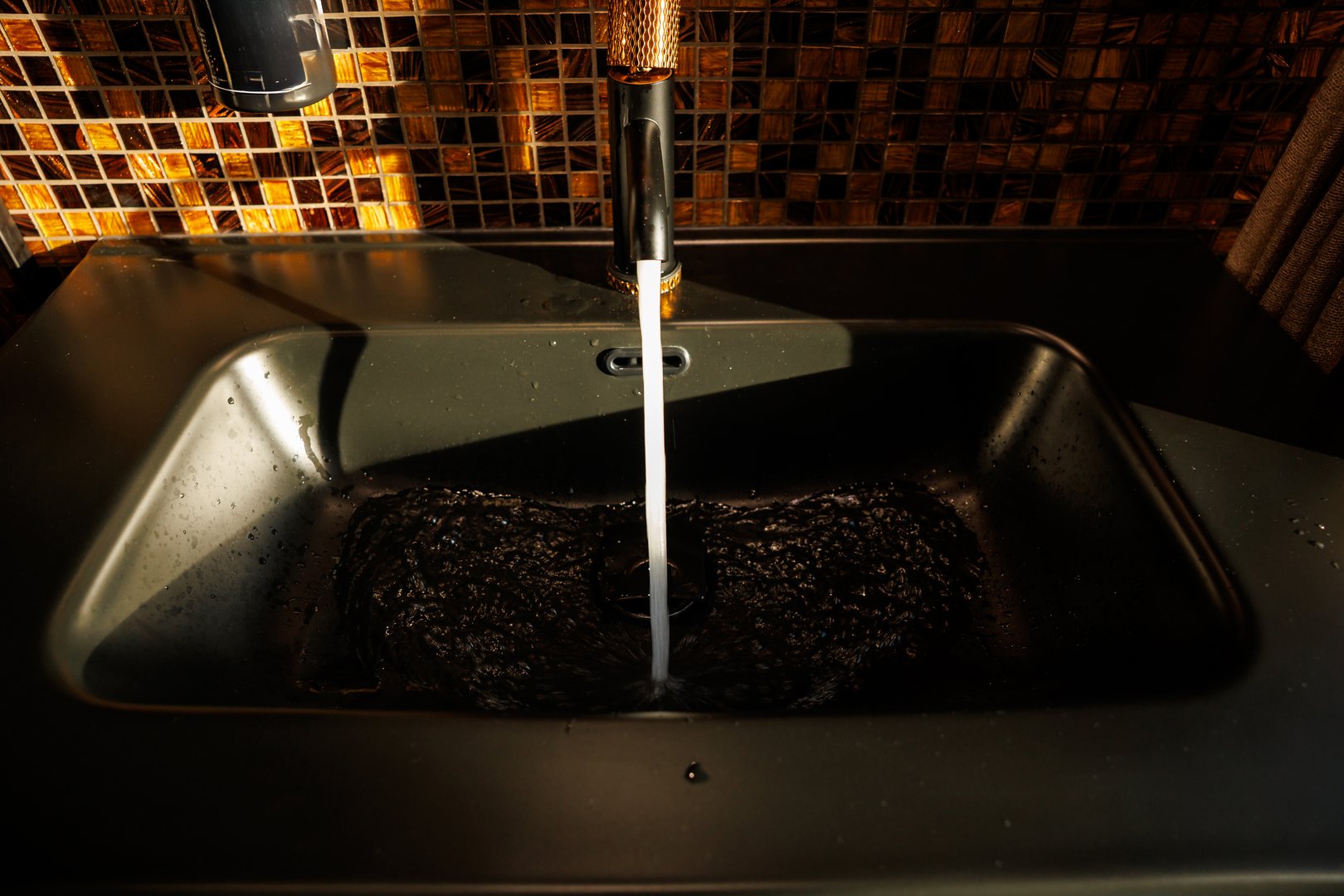 A black sink with water flowing from a metallic faucet with a copper colored handle, set against a mosaic tile wall in warm brown and gold tones.