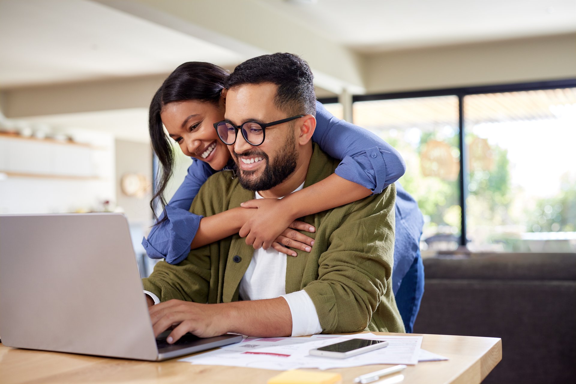 Young happy multiethnic couple having fun while using laptop at home. Loving young wife embracing husband from behind while typing on laptop. Happy couple making to do list of purchases and discussing future plans.