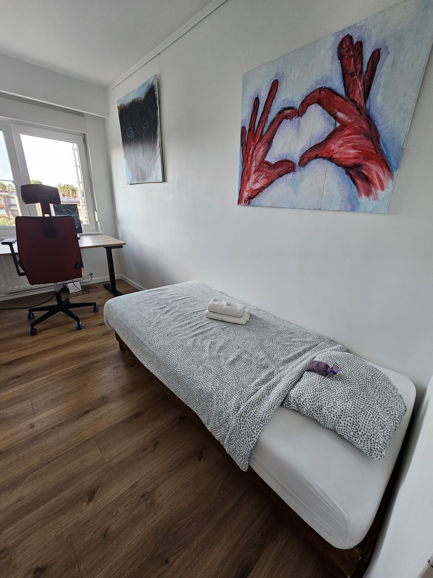 Minimalist room with a single bed, desk chair, and artworks on walls. Bed has a folded towel and small purple item on it.