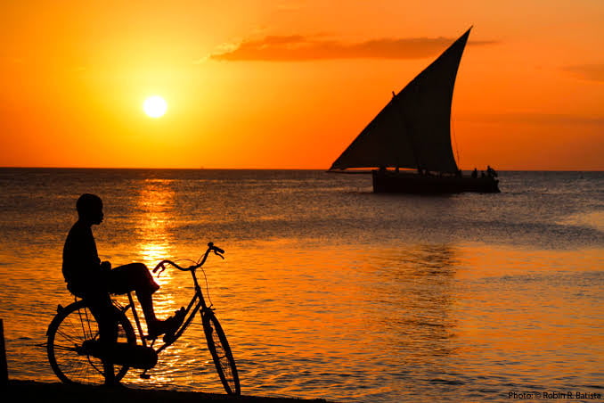 Silhouette of a person on a bicycle at sunset by the sea, with a sailboat on the horizon.