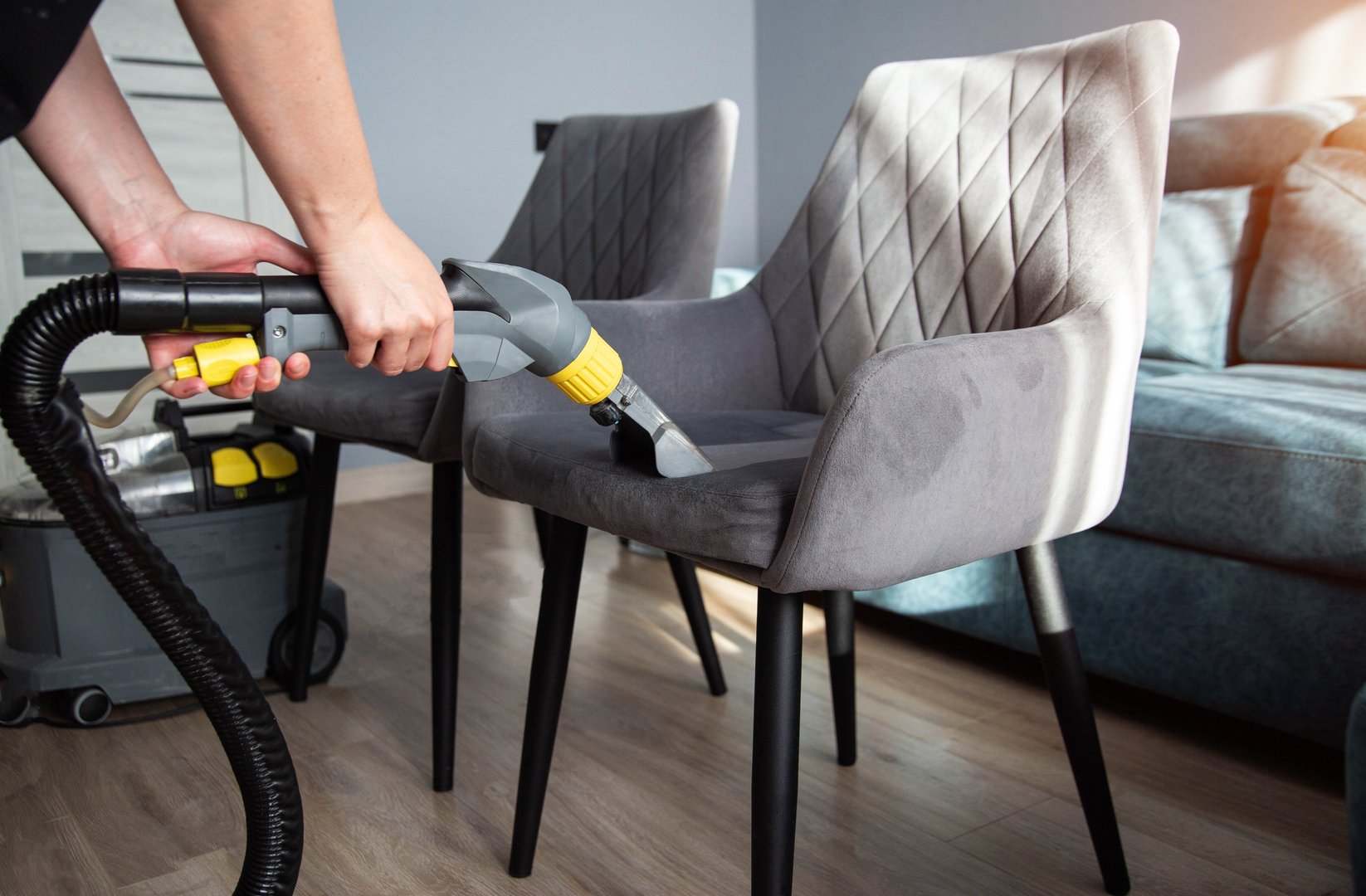 A person is cleaning a gray upholstered chair using a steam cleaner in a bright living room. Sunlight floods the space, highlighting the modern furniture and decor.