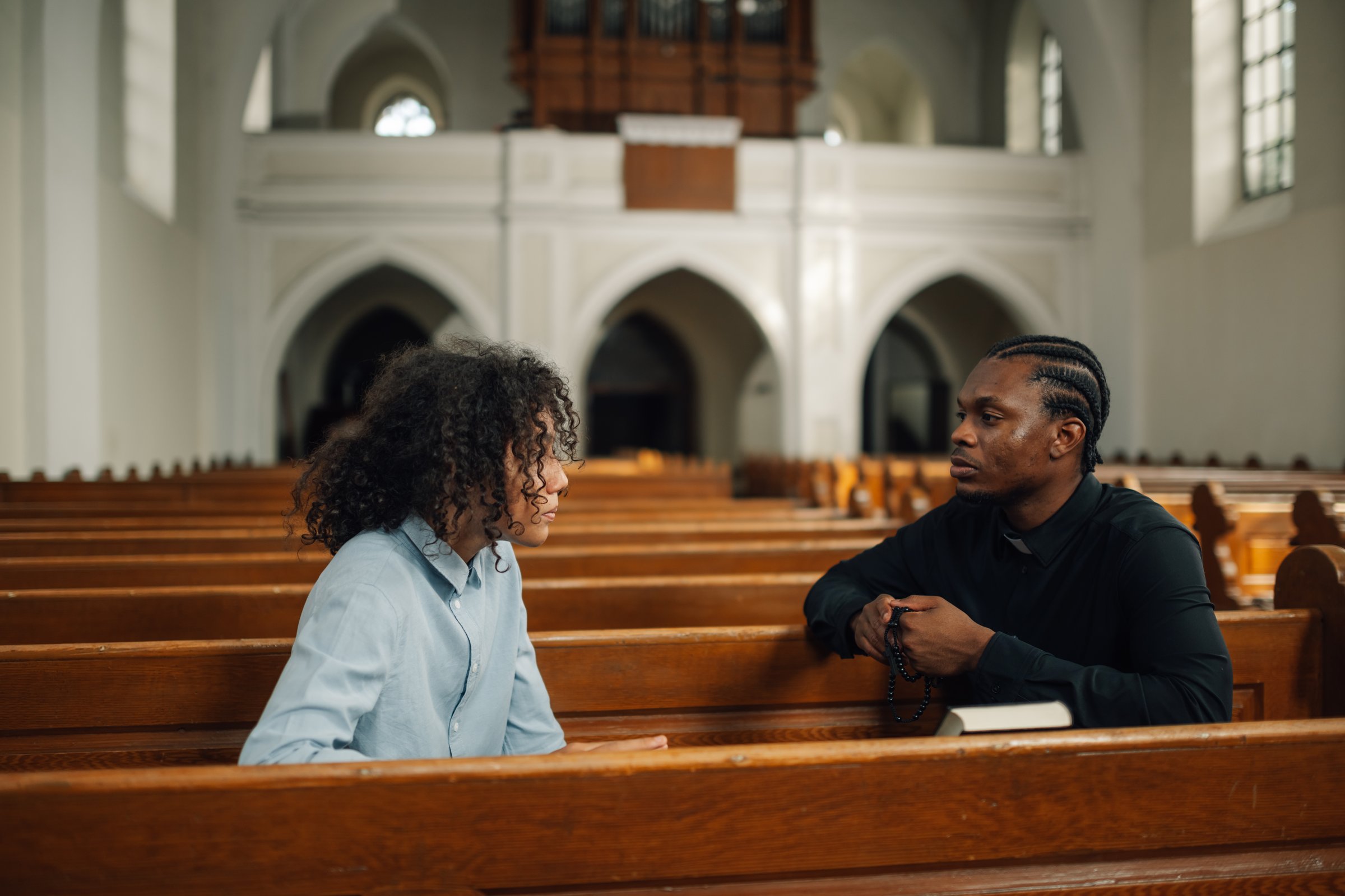 Young man talks to a priest in a church, seeking guidance and support. The priest listens attentively, holding rosary beads. The scene conveys faith, hope, and understanding in a place of worship