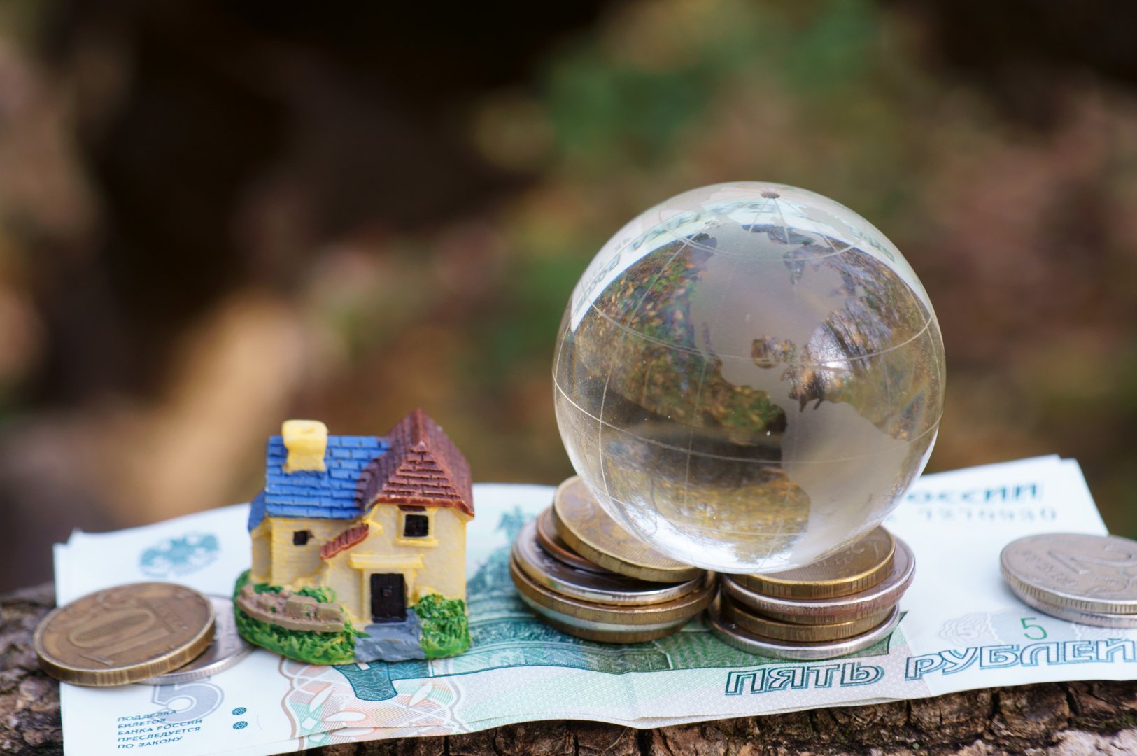 Glass transparent globe with coins in close-up. There is a toy house nearby.