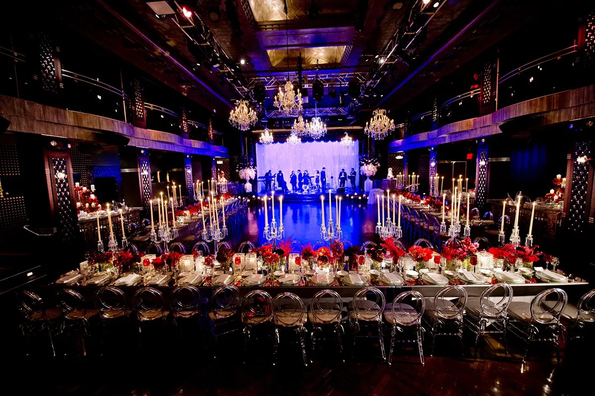Elegant banquet hall with long tables, red floral centerpieces, tall candelabras, and a stage with musicians under chandeliers.