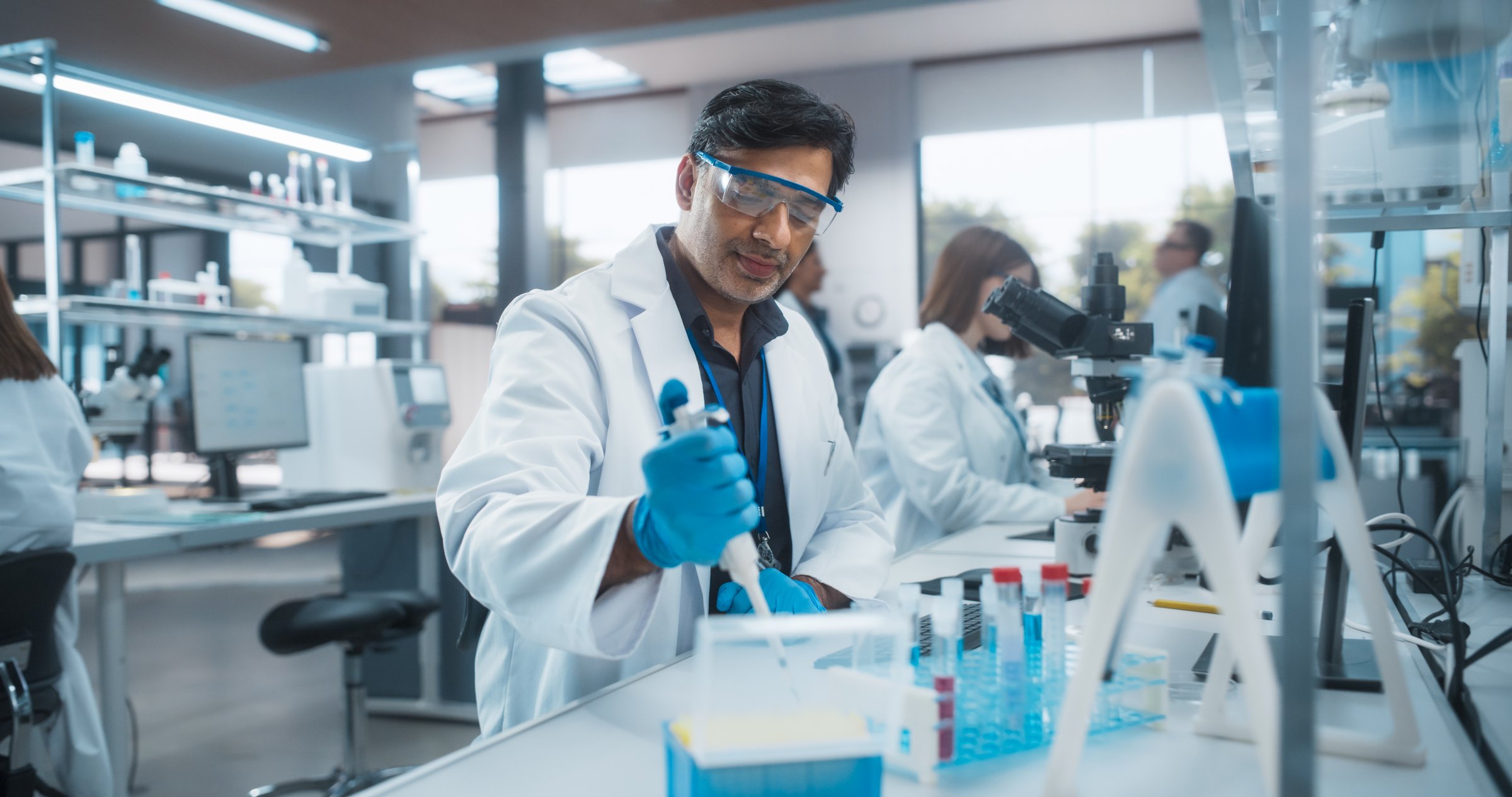 Indian Male Research Scientist Using Micropipette to Mix Liquids in a Test Tube in a Modern Laboratory. Scientists are Conducting Research with the Help of Microscopes and Desktop Computers
