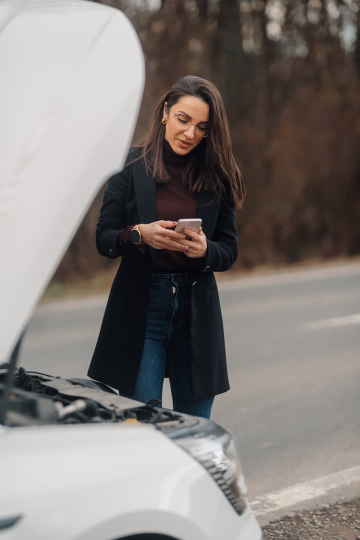 Elegant businesswoman using mobile phone, standing near broken car with open hood, calling for help