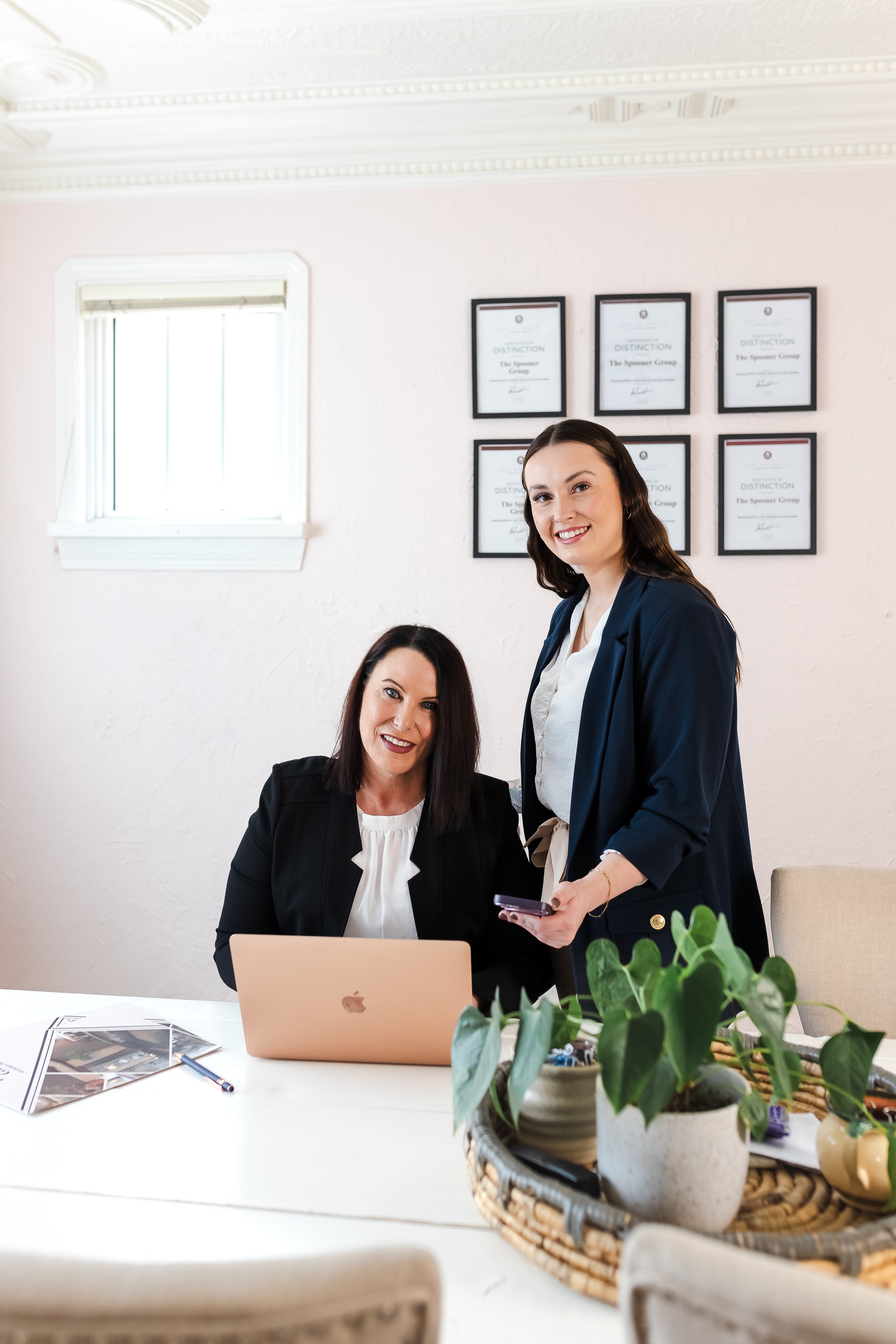 Two women in business attire smiling in an office, with one seated at a laptop and the other standing, holding a phone.