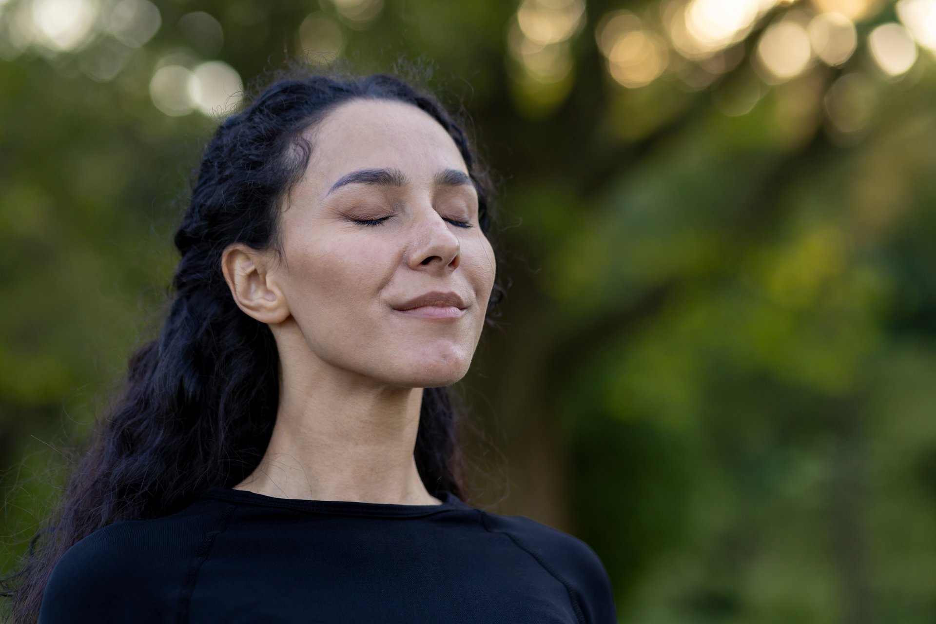A serene Hispanic woman closes her eyes, enjoying a tranquil moment outdoors surrounded by greenery, feeling calm and peaceful.