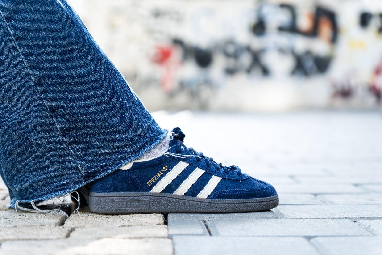 Tel Aviv, Israel - February 14, 2025.Stylish close-up of feet of a person wearing blue Adidas Spezial sneakers and frayed denim jeans, standing on a paved urban surface with graffiti background.