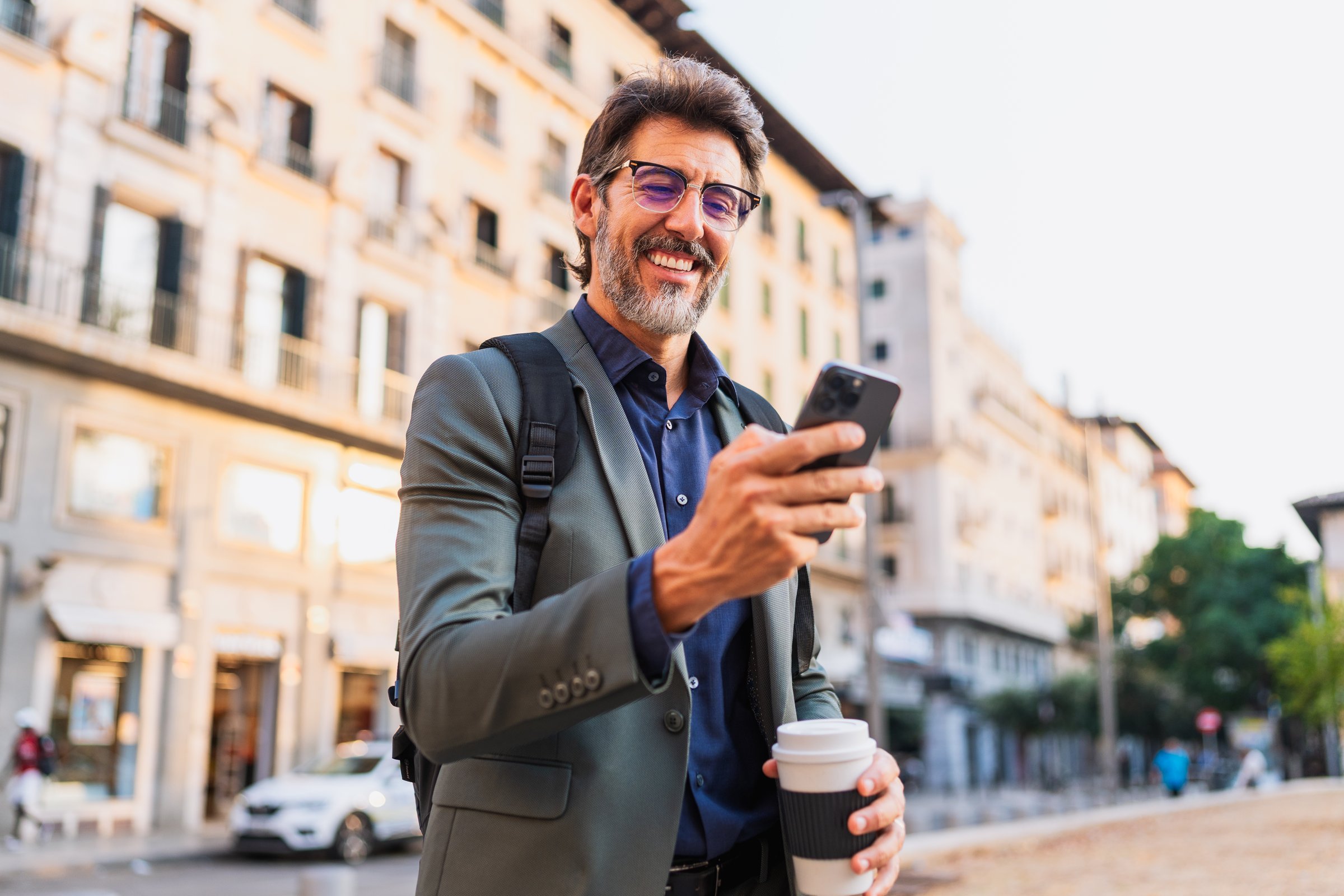 Confident professional man smiling while holding coffee and checking his phone on a city street. Concept of modern business lifestyle.