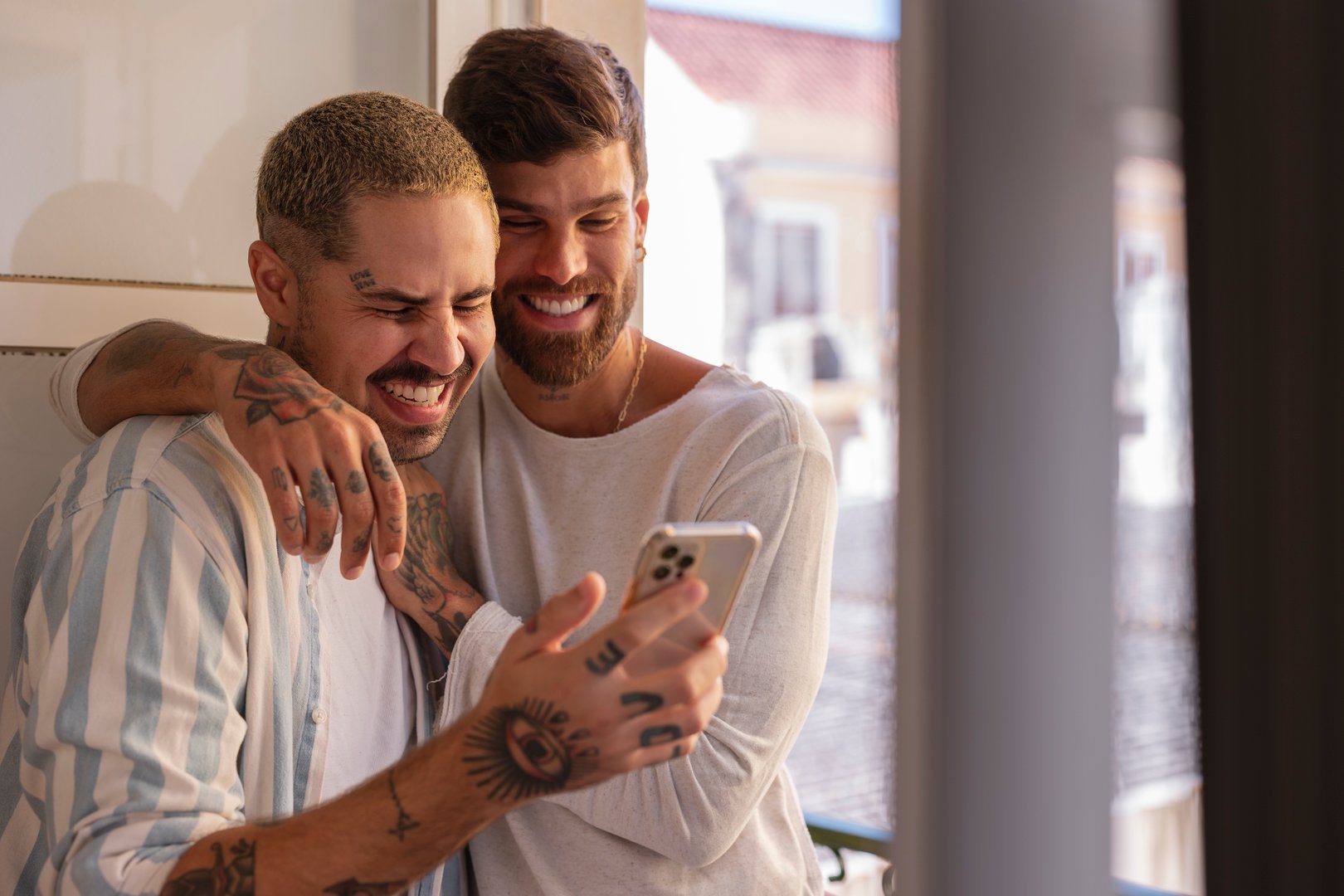 Two men embracing and laughing while using a smartphone by the window in their home, enjoying a moment of connection and happiness together