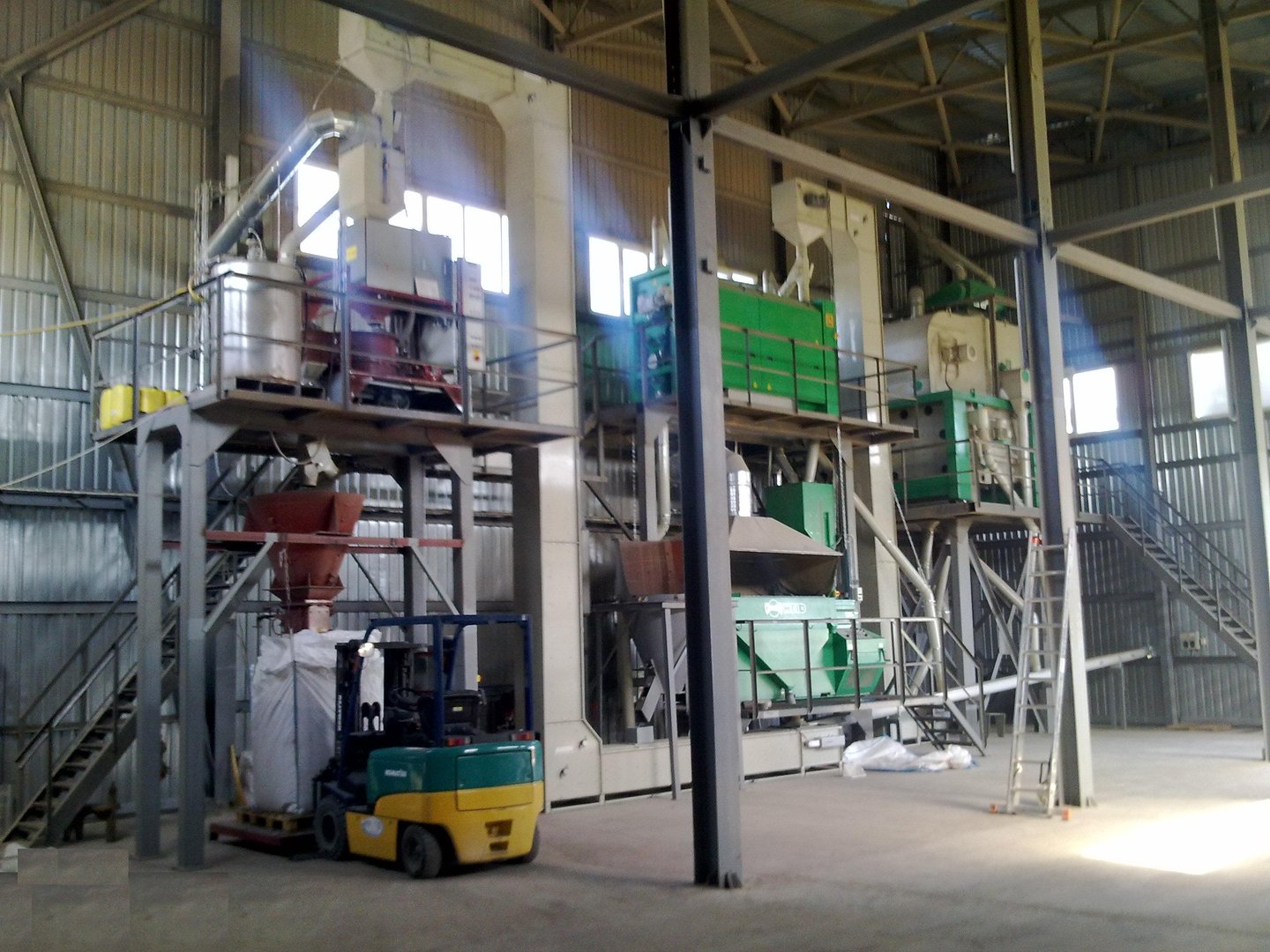 Industrial facility interior with green metal structures, large machines, pipes, and a staircase leading to an upper level.